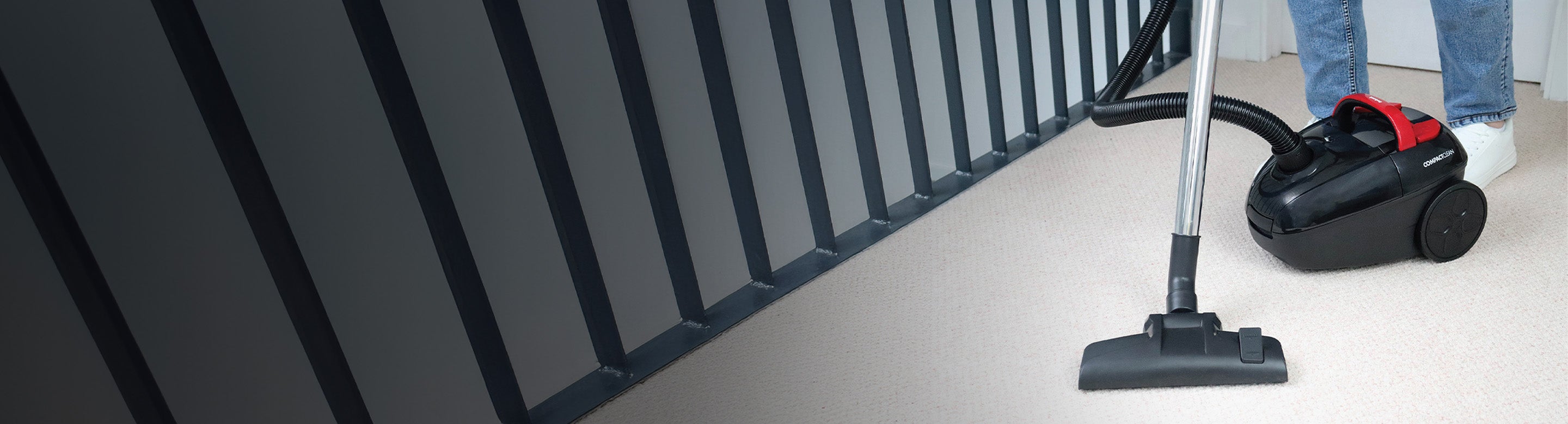 A person using a black and red canister vacuum cleaner on a light-colored carpet near a dark railing, with only their lower legs and white shoes visible.