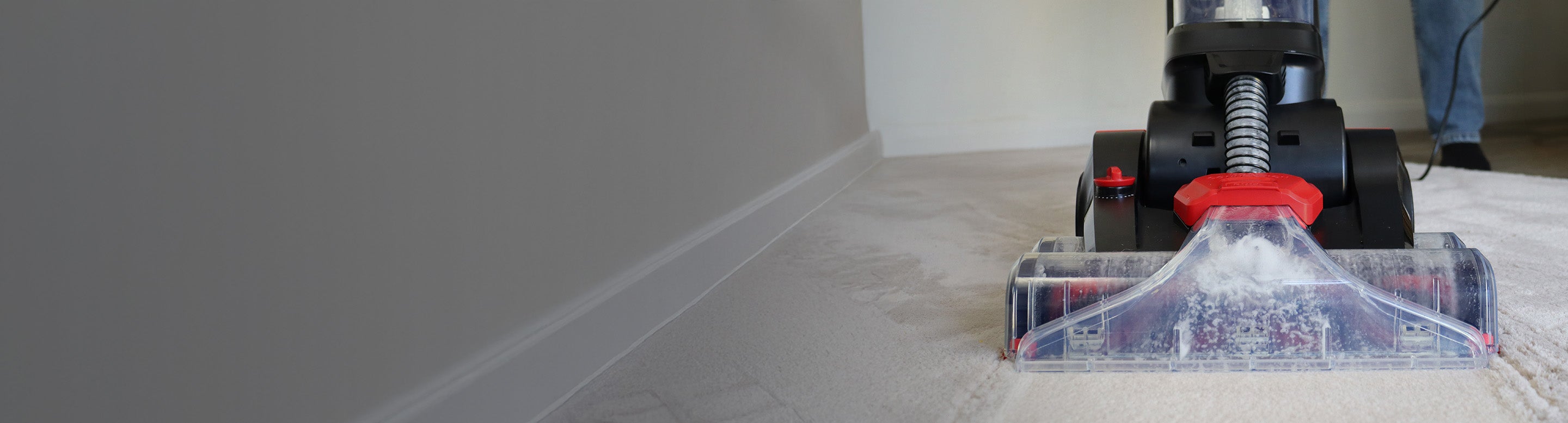 Close-up of a carpet cleaning machine being pushed over a light-colored carpet, with a person’s legs and shoes partially visible in the background on the right.