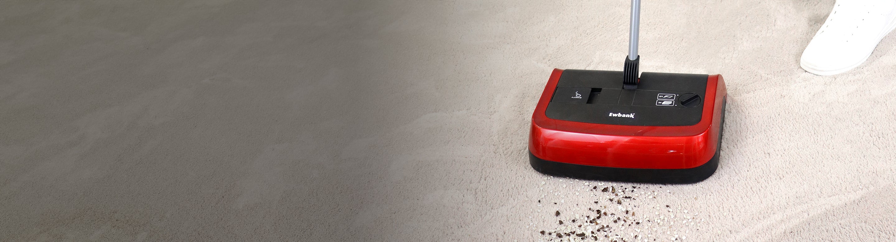 A red and black manual floor sweeper is being pushed across a light-colored floor, picking up scattered debris; part of a white shoe is visible in the top right corner.