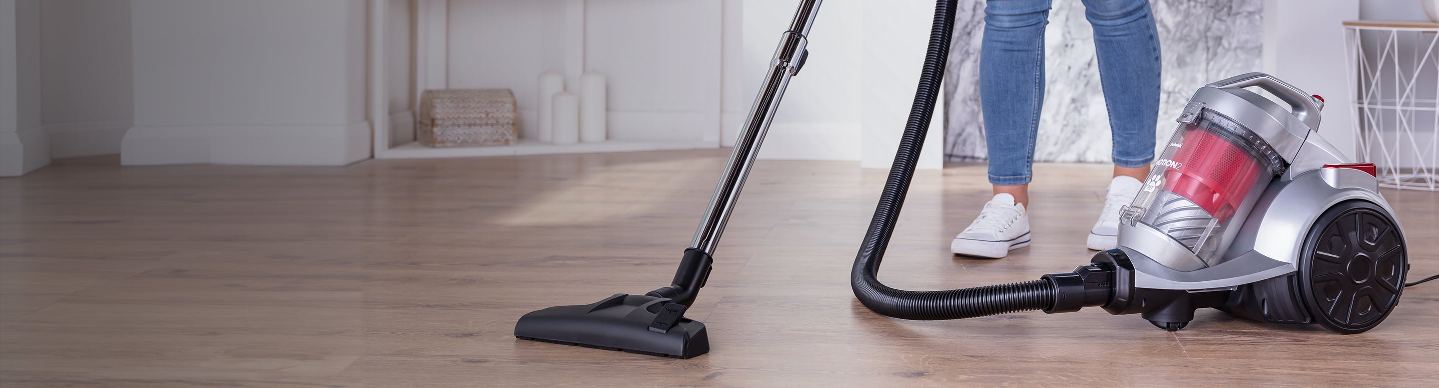 Person vacuuming a clean, light-colored hardwood floor with a modern canister vacuum cleaner; only their legs and casual shoes are visible in the bright, tidy room.