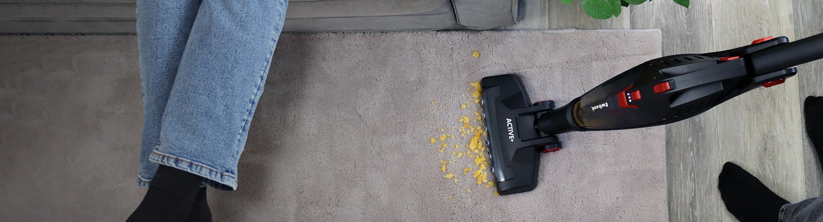A person vacuums crumbs from a beige carpet next to a gray sofa, wearing blue jeans and black socks. The vacuum cleaner is picking up yellow debris from the carpet.