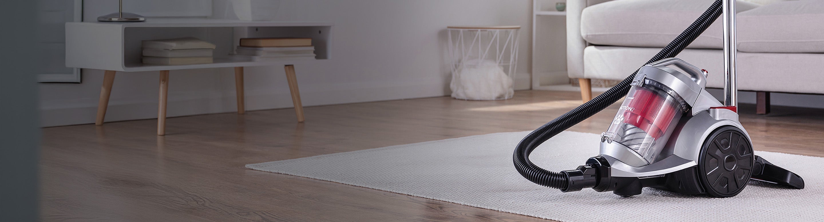 A canister vacuum cleaner with a hose sits on a light-colored rug in a modern living room with wooden floors and minimalist furniture in the background.