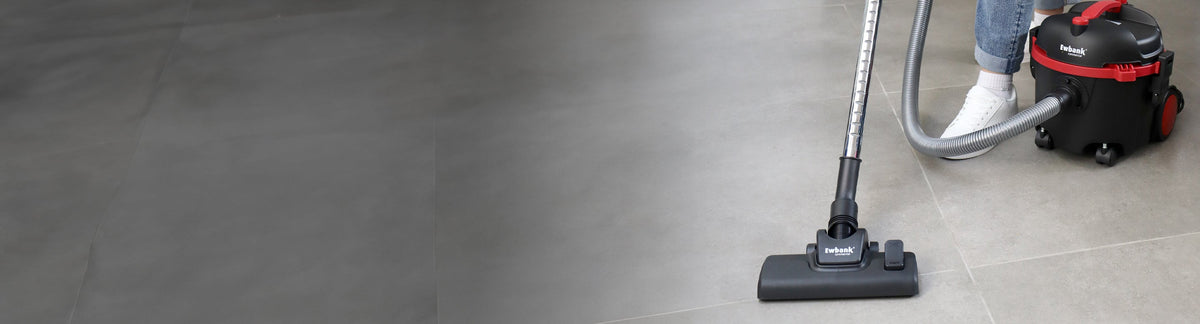 A person vacuuming a tiled floor with a canister vacuum cleaner; only their lower legs and feet are visible, and the vacuum is predominantly black with red accents.