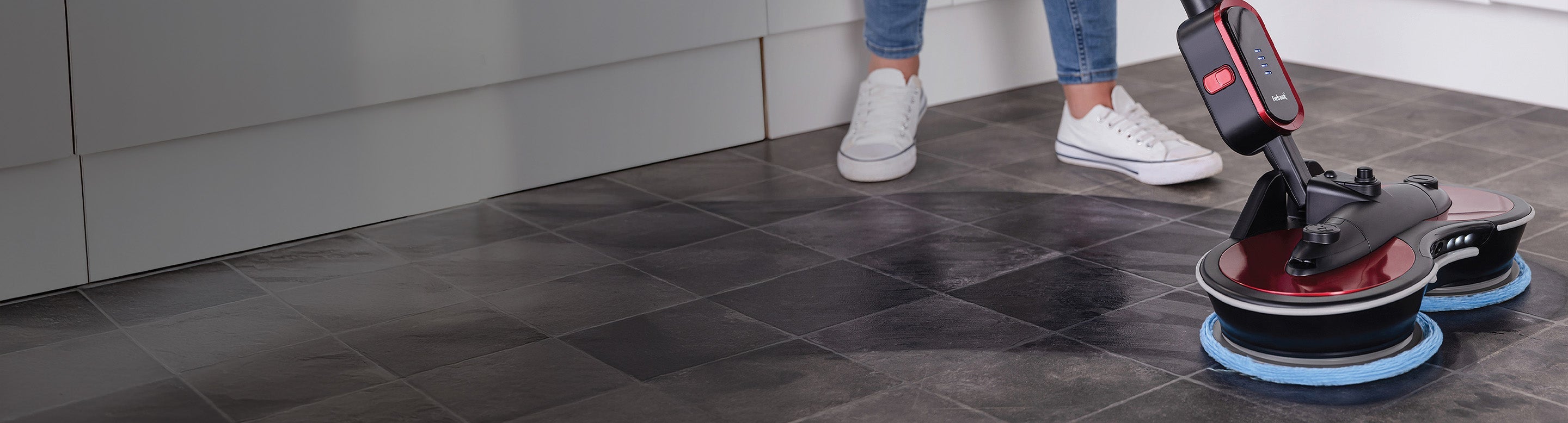 A person uses an electric floor cleaning machine with dual rotating pads to clean a tiled kitchen floor. Only the lower legs and feet of the person are visible.
