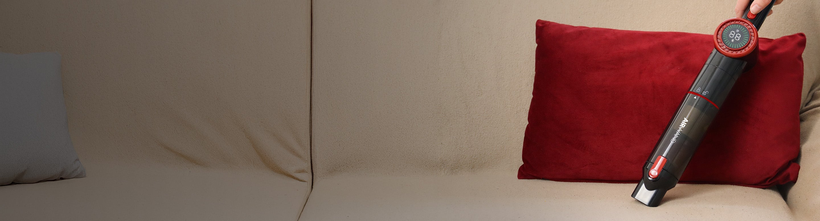 A person uses a handheld vacuum cleaner with red accents to clean a red pillow on a beige sofa, with a light blue pillow also visible.