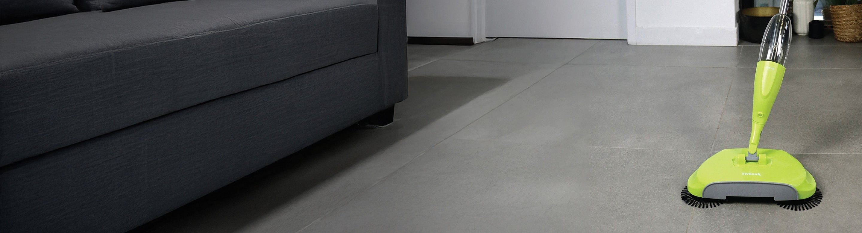 A green floor-cleaning machine is on a gray tile floor near a dark gray sofa in a modern living room. White walls and furniture are visible in the background.