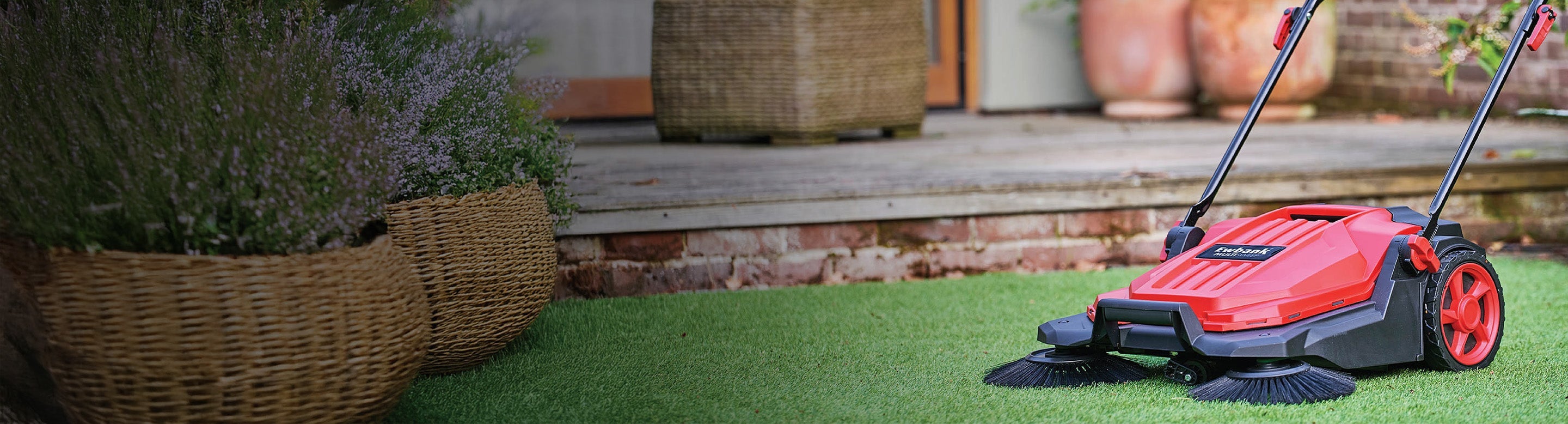 A red and black push lawn sweeper with two rotating brushes sits on green artificial grass near large woven plant baskets and a wooden patio.