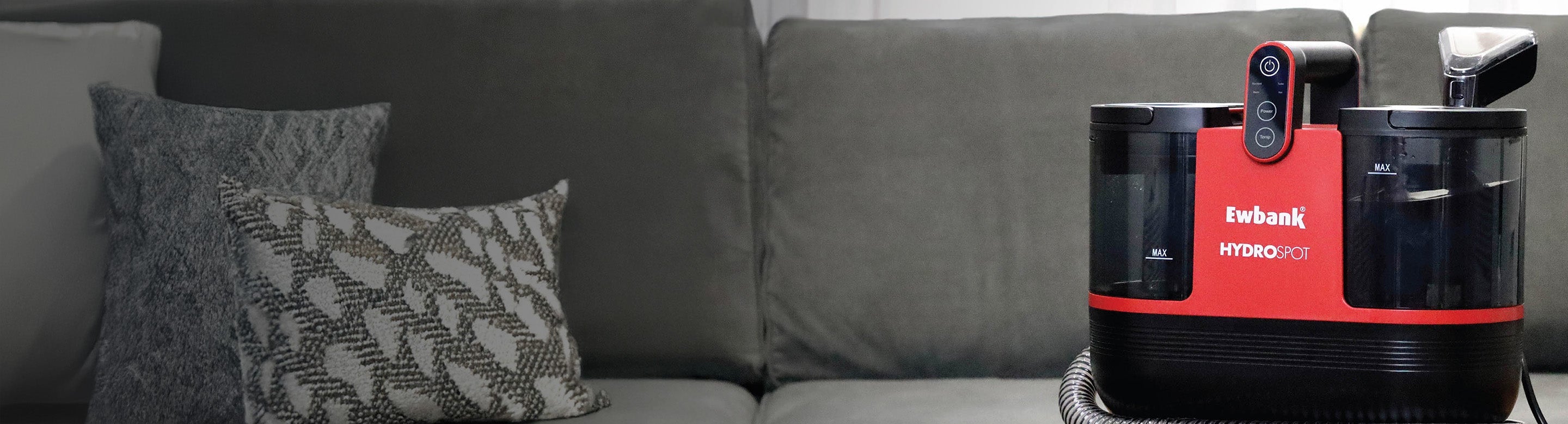 A red and black Ewbank Hydro Pet cleaning machine sits on the arm of a gray sofa with patterned gray-and-white cushions in the background.
