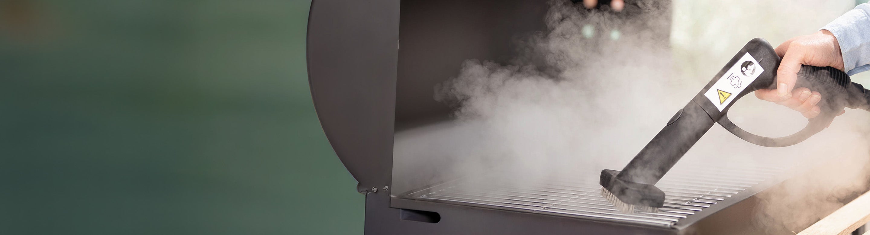 A person uses a handheld steam cleaner to clean the metal grates of a barbecue grill, with visible steam rising from the surface.