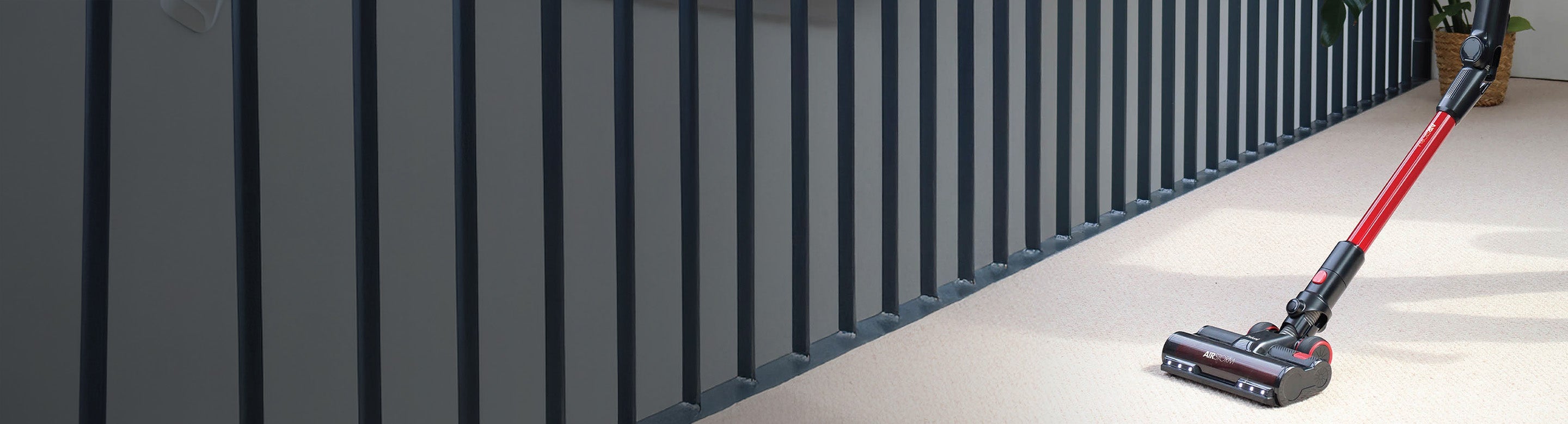 A cordless vacuum cleaner with a red handle is cleaning a light-colored carpet next to a modern, dark gray railing.