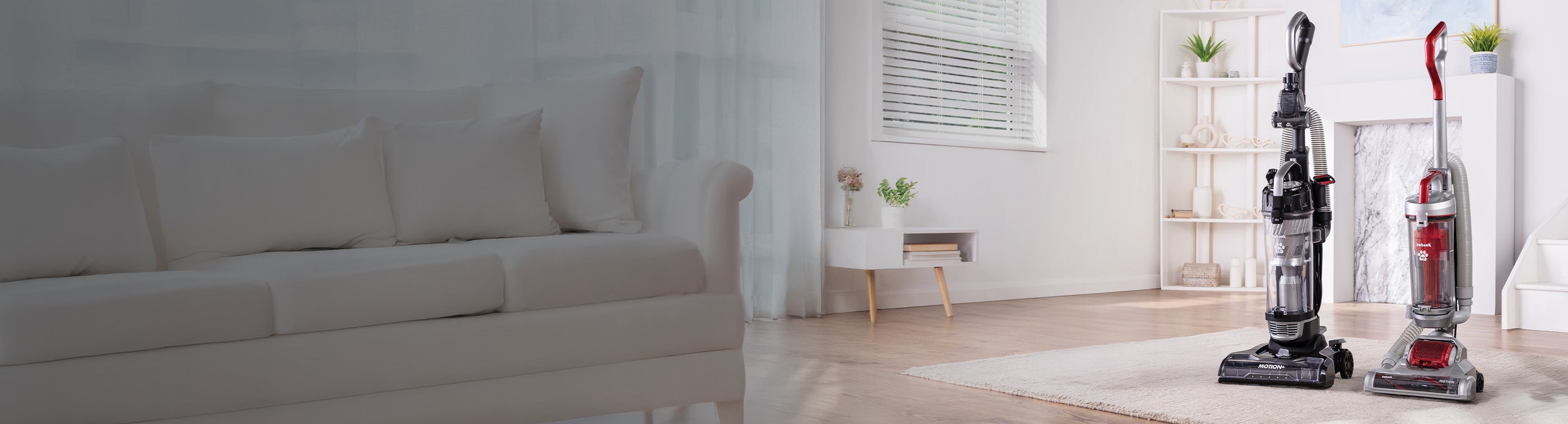 Two upright vacuum cleaners stand on a light wooden floor in a bright, modern living room with a white sofa, side table, and shelves in the background.