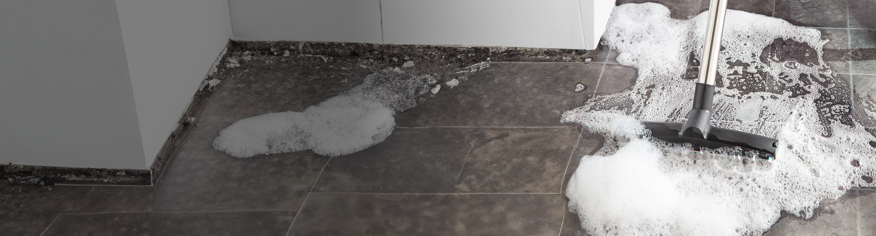 A mop pushes soapy water and foam across a dark tiled floor, cleaning near the corner of a room with baseboards.