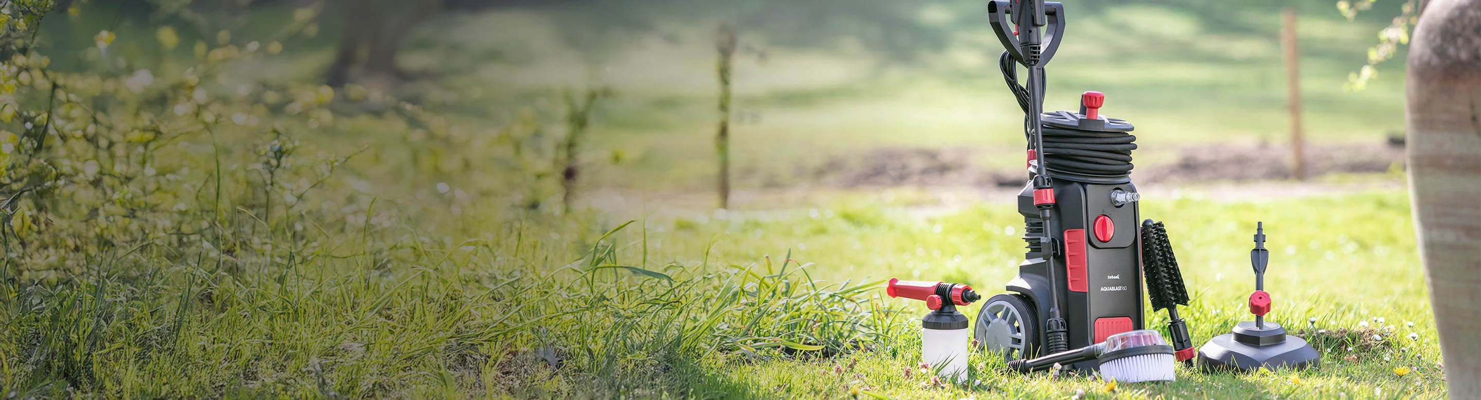A black and red pressure washer with various attachments sits on green grass outdoors, with a blurred garden and trees in the background.