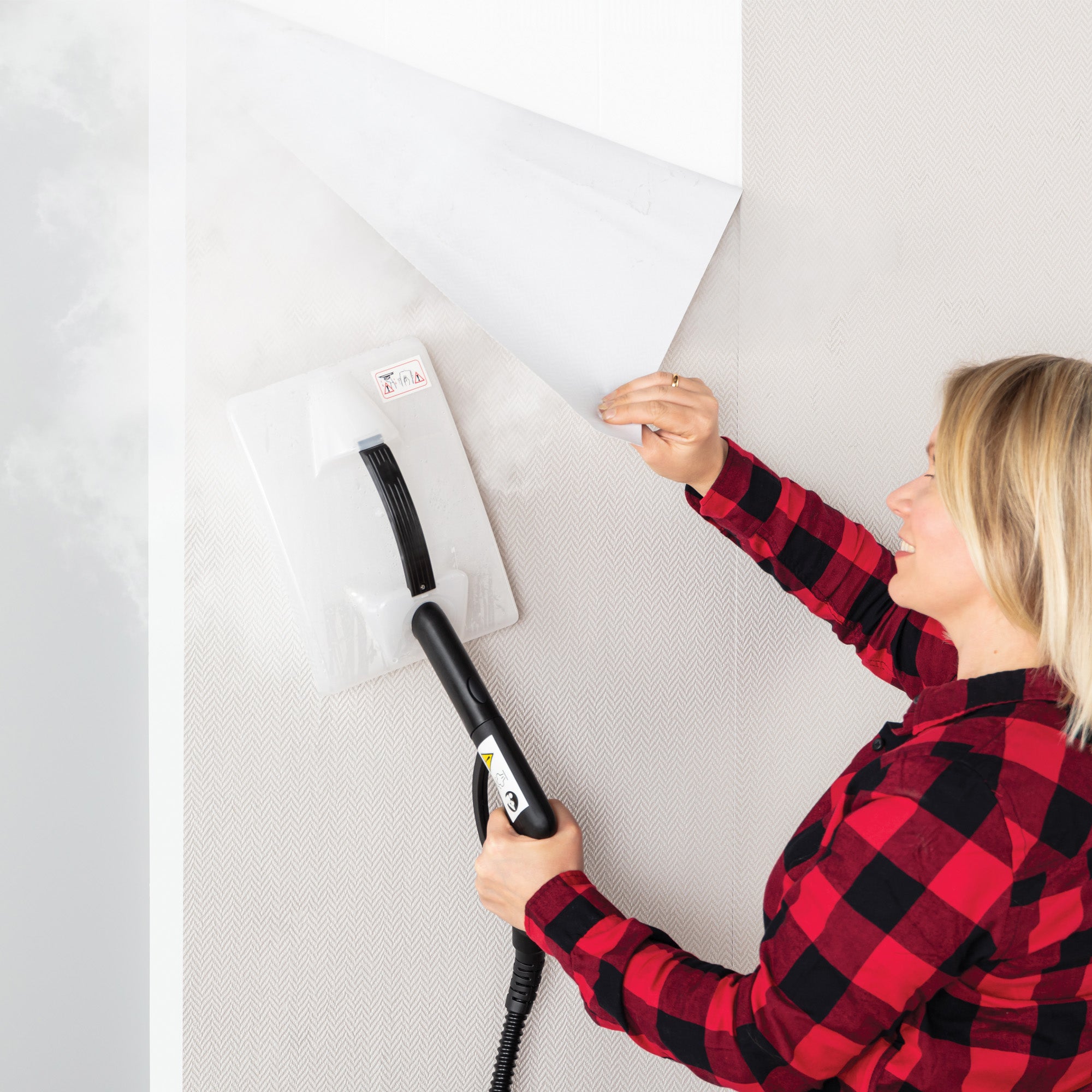 Wearing a red plaid shirt, a person uses the STEAMDYNAMO+ 2000W Steam Cleaner & Sanitiser to peel wallpaper, holding the steamer pad with one hand and lifting the wallpaper with the other for effective, chemical-free cleaning.