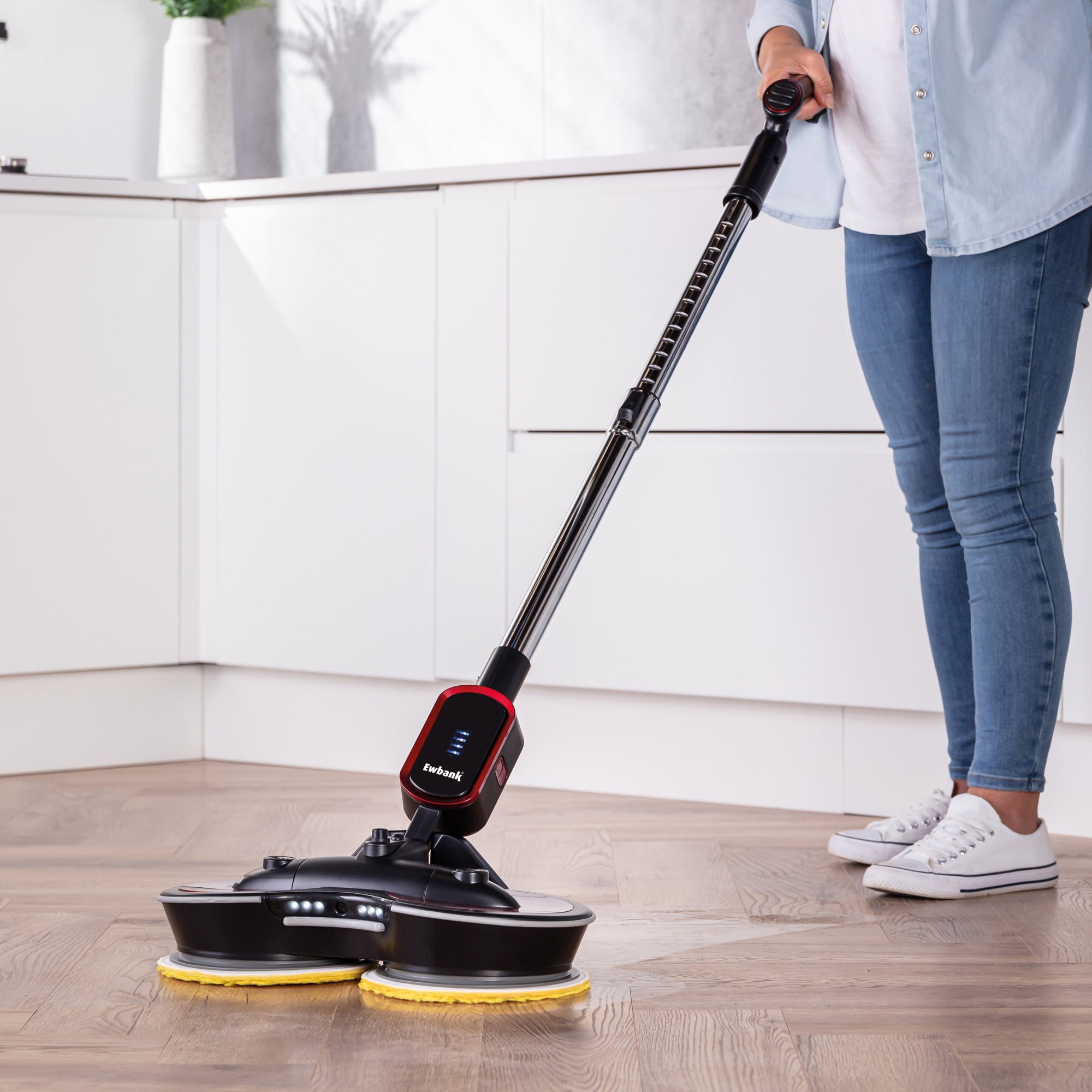 A person cleans a wooden floor in a modern, white kitchen using the FP90 Lightweight Cordless Floor Polisher & Cleaner with yellow pads. Only the lower half of the person is shown.