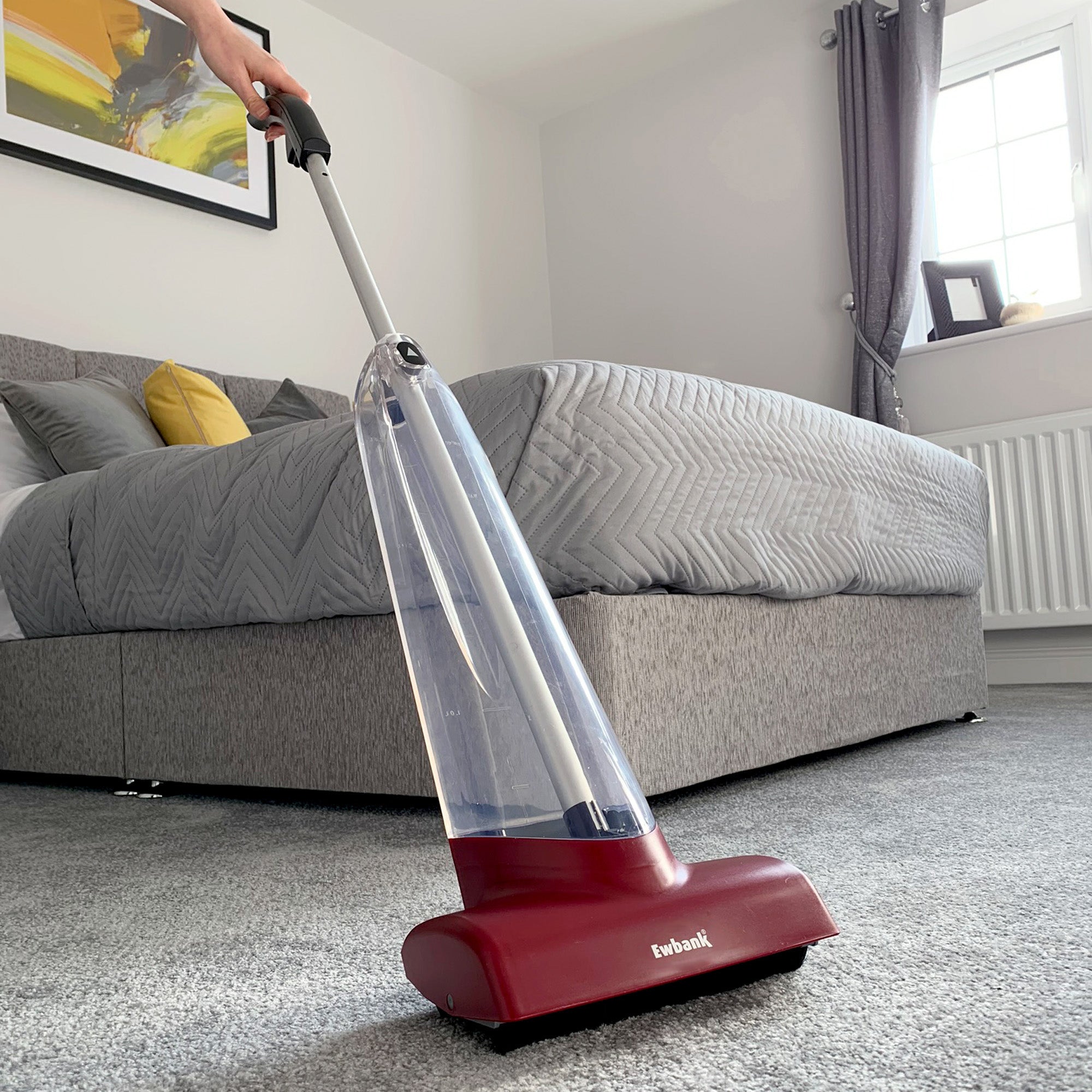 A person uses a red Cascade Manual Carpet Shampooer by Ewbank on a bedroom carpet. The modern room includes a large gray bed with cushions, geometric bedding, gray curtains, and a sunny window.