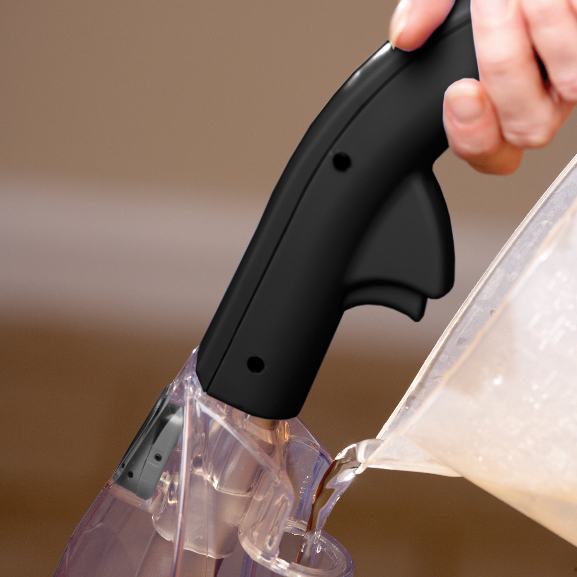 A hand holds the black handle of the Cascade Manual Carpet Shampooer by Ewbank in red, with another hand pouring carpet shampoo into its clear container. The background is neutral and out of focus.