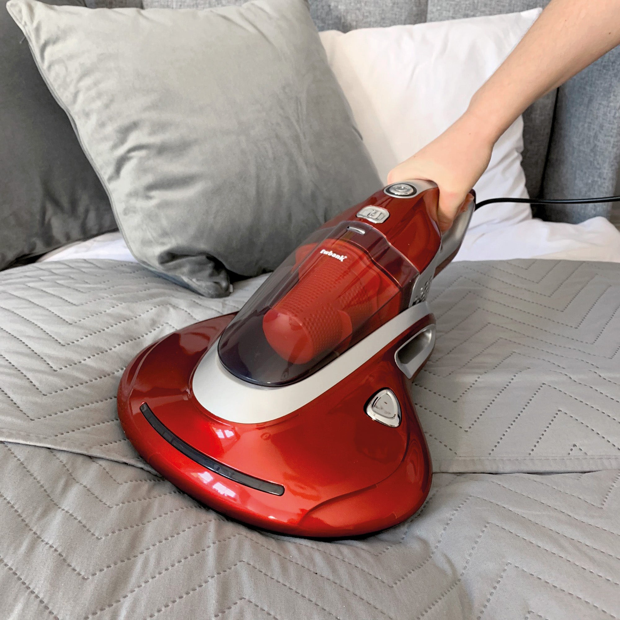 Red vacuum cleaner on a bed with a person's hand holding it