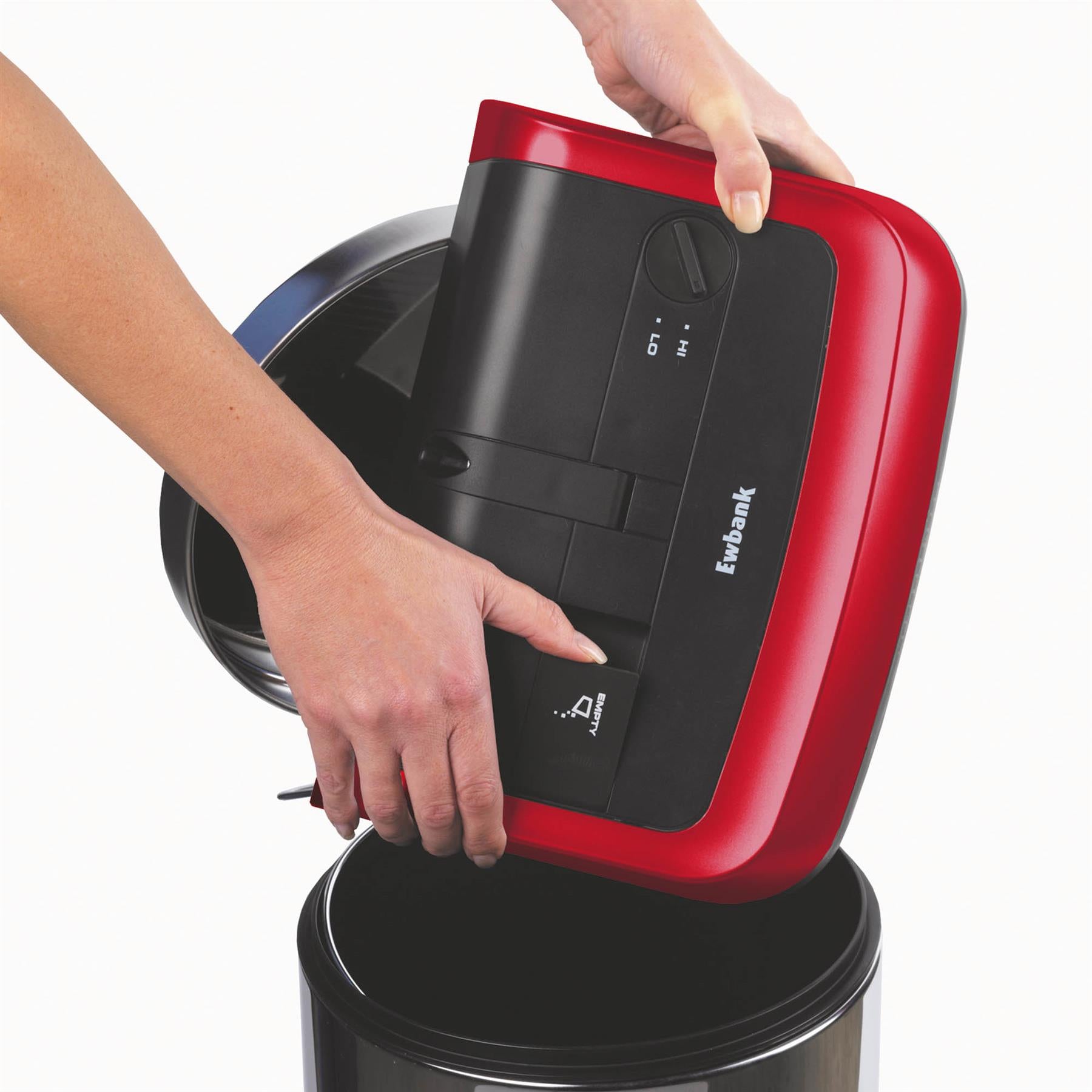 A person uses the Evo3 Manual Carpet Sweeper, holding its red and black body over a black trash can to empty shredded paper, making carpet cleaning easy and hygienic.