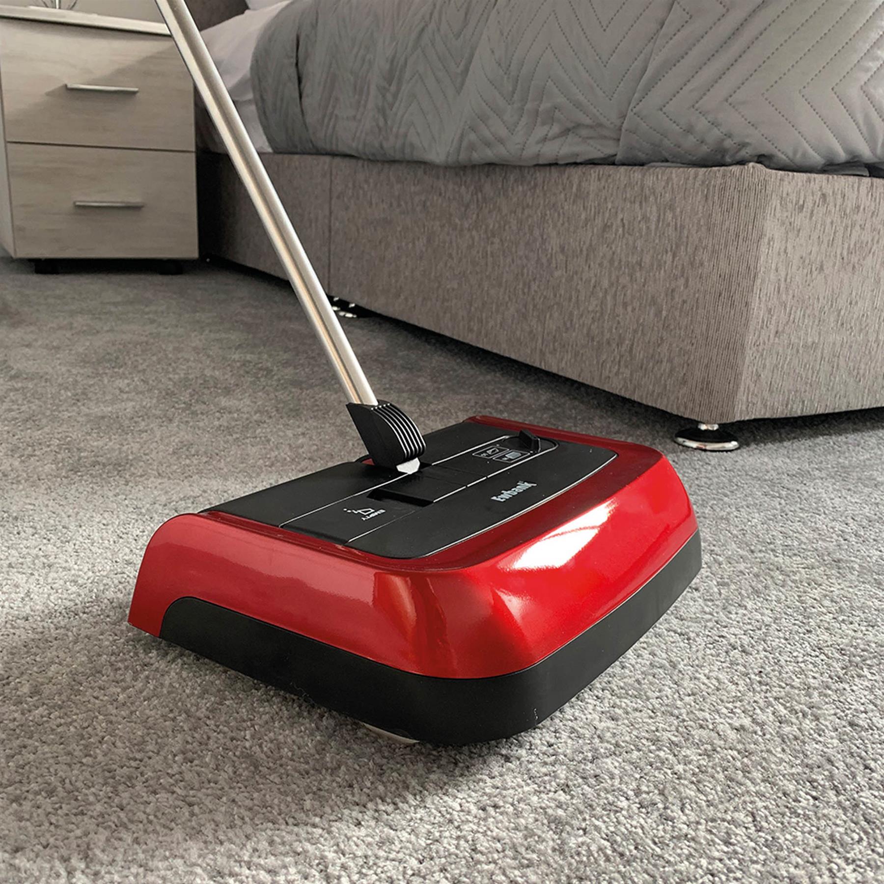 The Evo3 Manual Carpet Sweeper, featuring a red design, adjustable brush height, and metal handle, is used for hygienic carpet cleaning on a gray carpeted floor beside a bed and nightstand in a bedroom.