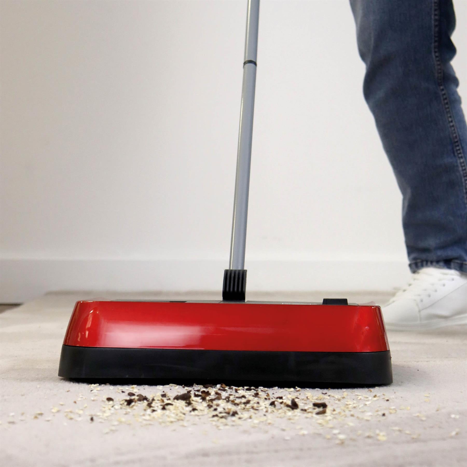 A person uses the Evo3 Manual Carpet Sweeper with adjustable brush height to clean debris on a light-colored carpet. Only their lower legs and white shoes are visible, highlighting efficient and hygienic carpet cleaning.