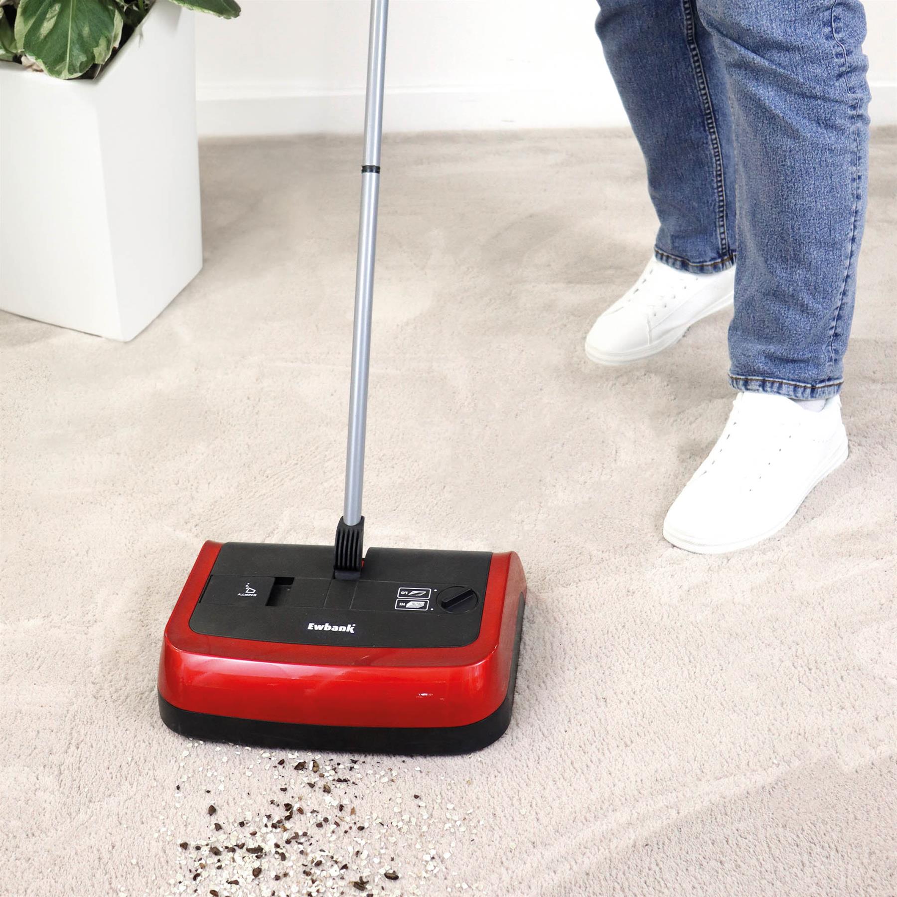Wearing blue jeans and white sneakers, a person uses the Evo3 Manual Carpet Sweeper—with adjustable brush height—to clean dirt from a light-colored carpet beside a green potted plant.