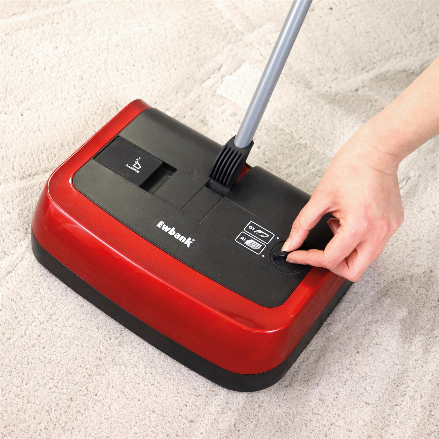 A person adjusts the brush height on a red Evo3 Manual Carpet Sweeper, ensuring hygienic cleaning on a light-colored carpet.