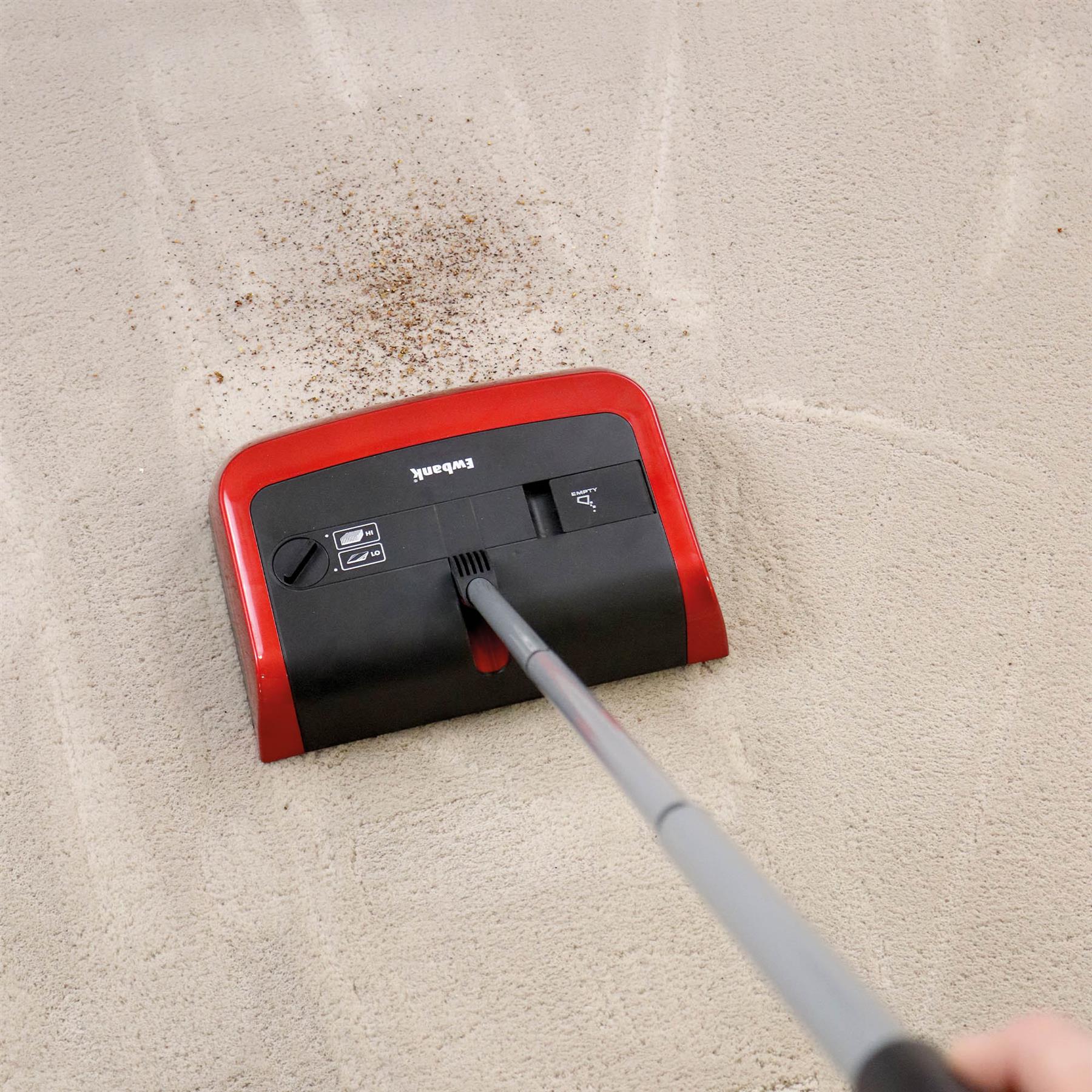 The Evo3 Manual Carpet Sweeper, featuring adjustable brush height and a red and black body, glides over a light-colored carpet, collecting dirt and leaving clean, parallel sweep lines for hygienic cleaning.
