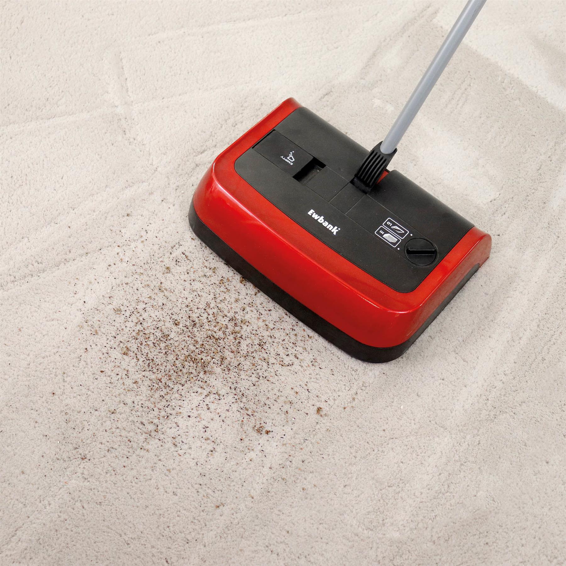 The Evo3 Manual Carpet Sweeper in red and black, with adjustable brush height, is used on a light-colored carpet to easily sweep up dirt and debris for a more hygienic clean.