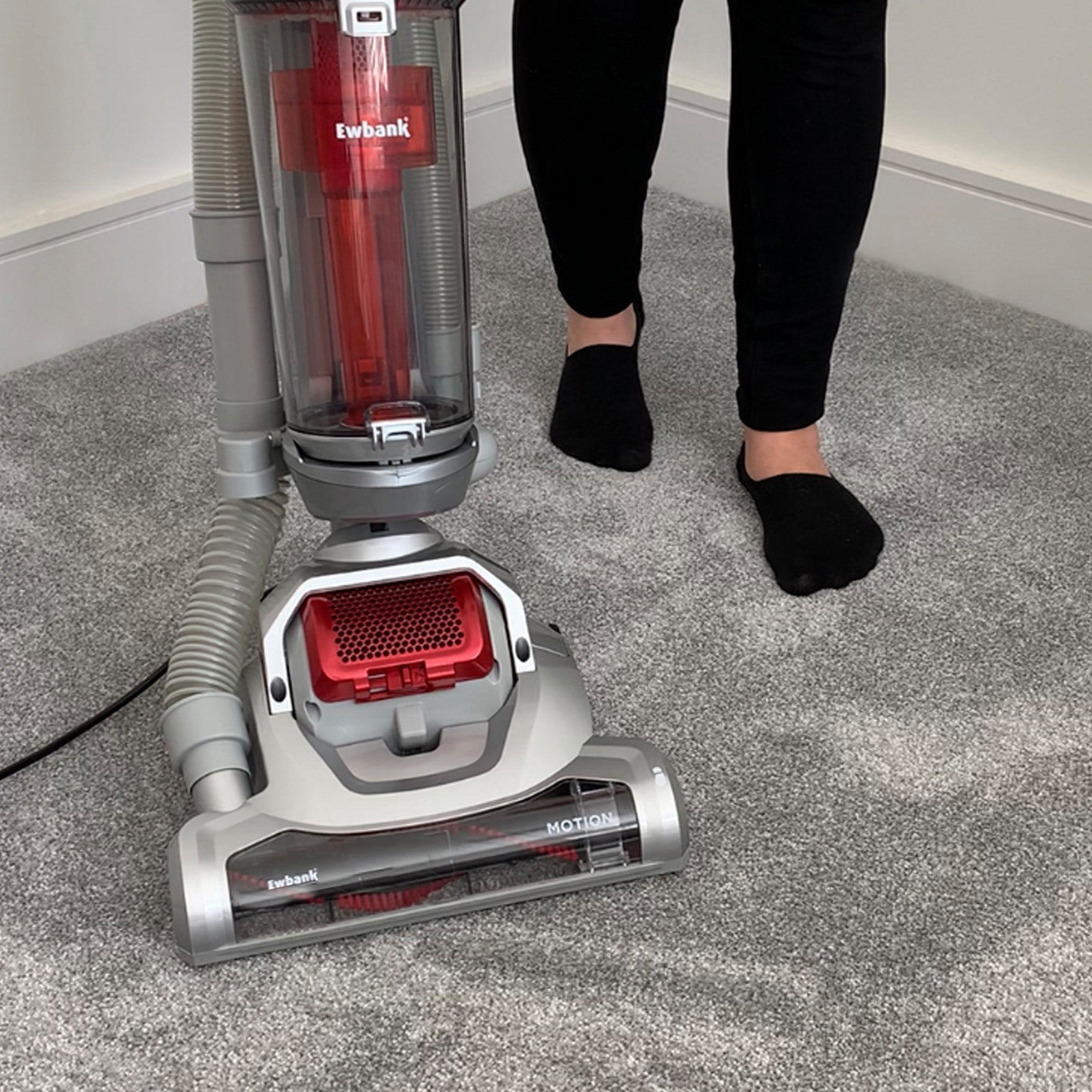 A person in black socks stands on a light grey carpet using a red and silver MOTION Pet Upright Bagless Vacuum Cleaner with cyclonic filtration, plugged in and near a white baseboard.
