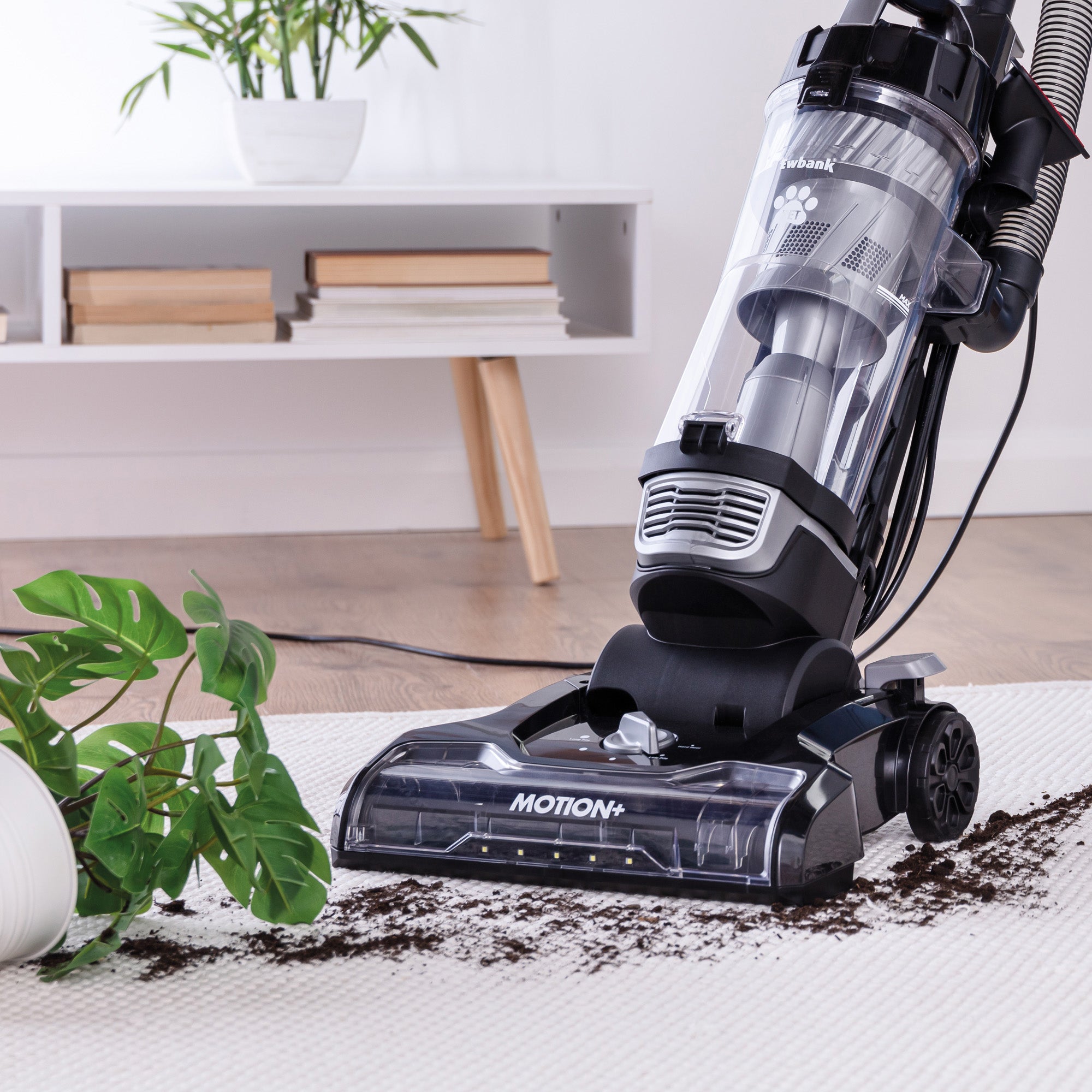 The MOTION+ Reach Pet Upright Bagless Vacuum Cleaner with multi-cyclone technology is cleaning spilled soil from a white carpet beside an overturned plant and pot in a modern living room.