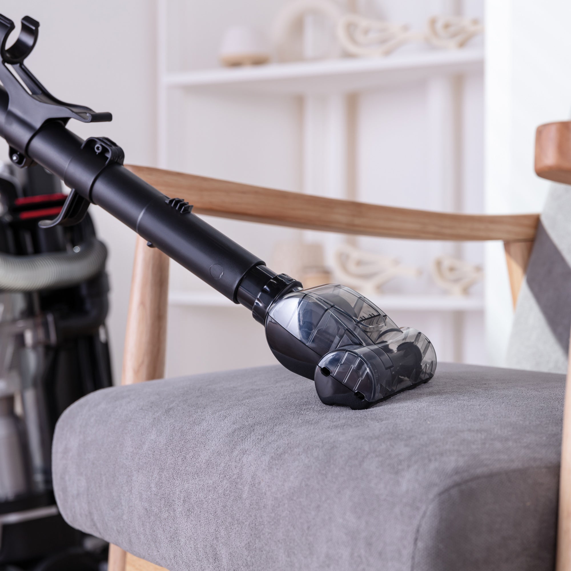 The MOTION+ Reach Pet Upright Bagless Vacuum Cleaner with a brush attachment is being used to deep clean a gray upholstered chair in a living room, with shelves and decorative items softly blurred in the background.