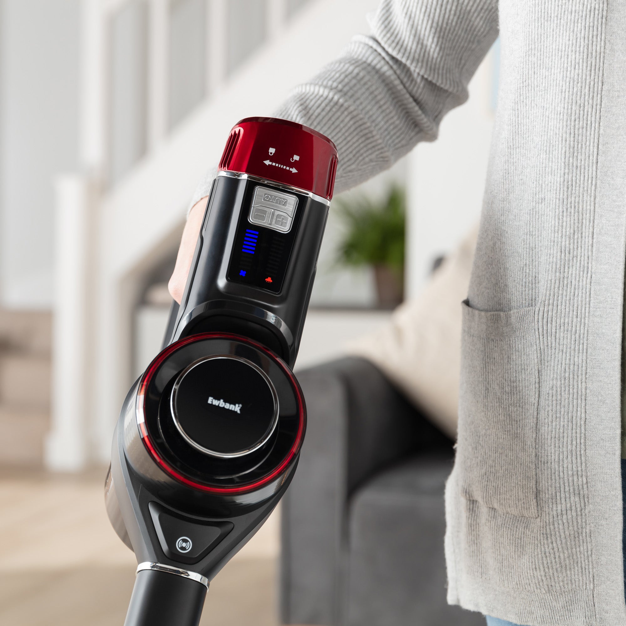 A person uses the AIRSTORM 2-in-1 Cordless Stick Vacuum Cleaner indoors; a staircase and gray sofa are visible in the background.