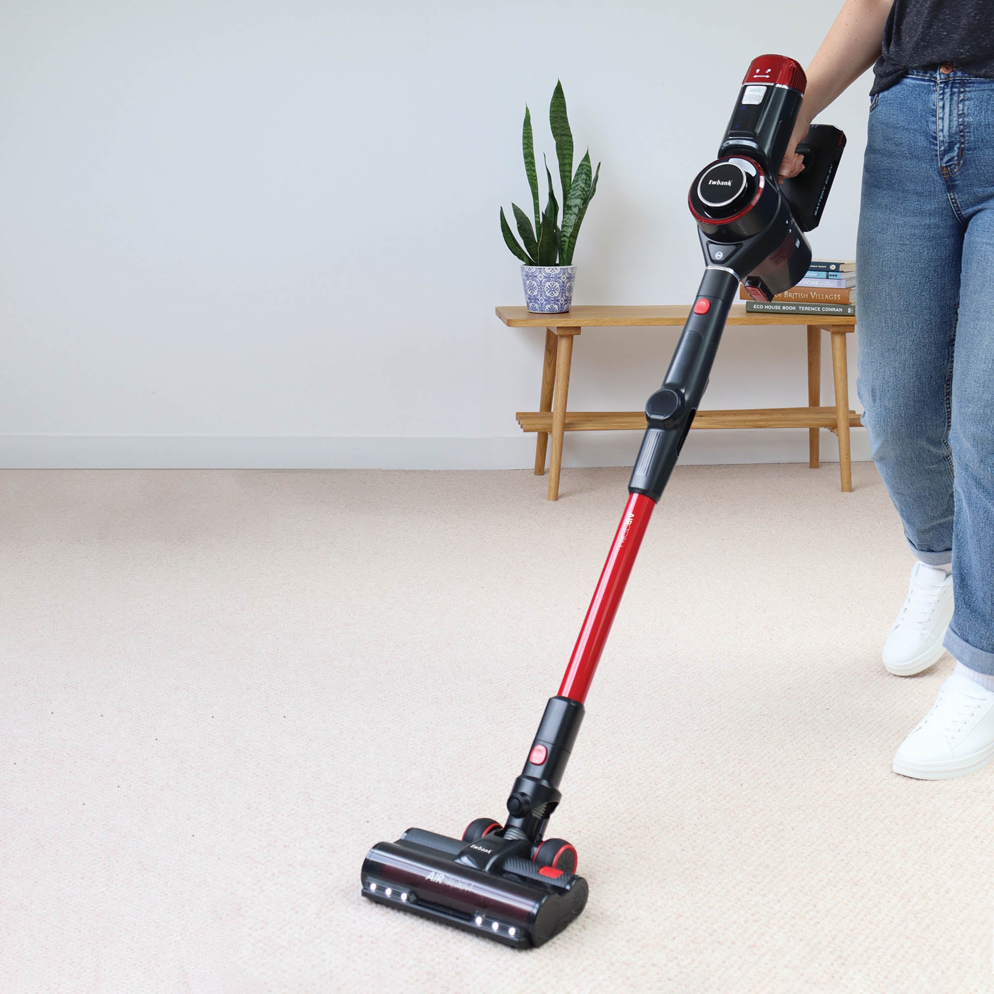 A person in jeans and white shoes vacuums a light-colored carpet using the red and black AIRSTORM 2-in-1 Cordless Stick Vacuum Cleaner. A wooden bench with a plant, books, and a candle is seen in the background.
