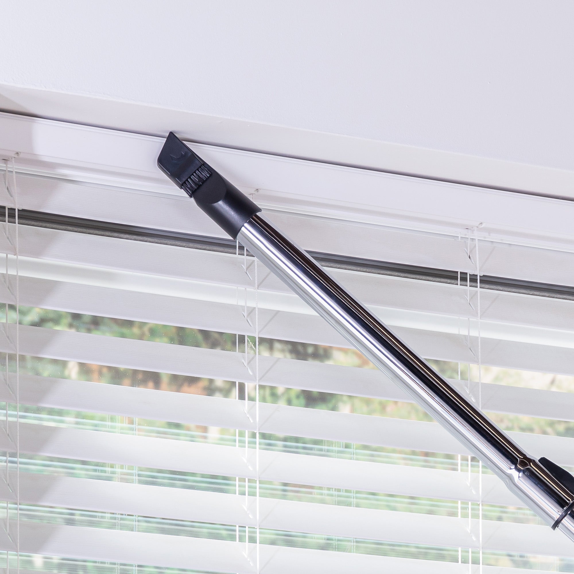 The Ewbank MOTIONLITE Bagless Cylinder Vacuum Cleaner, equipped with a narrow nozzle, is being used to clean the upper edge of a window frame above horizontal blinds.