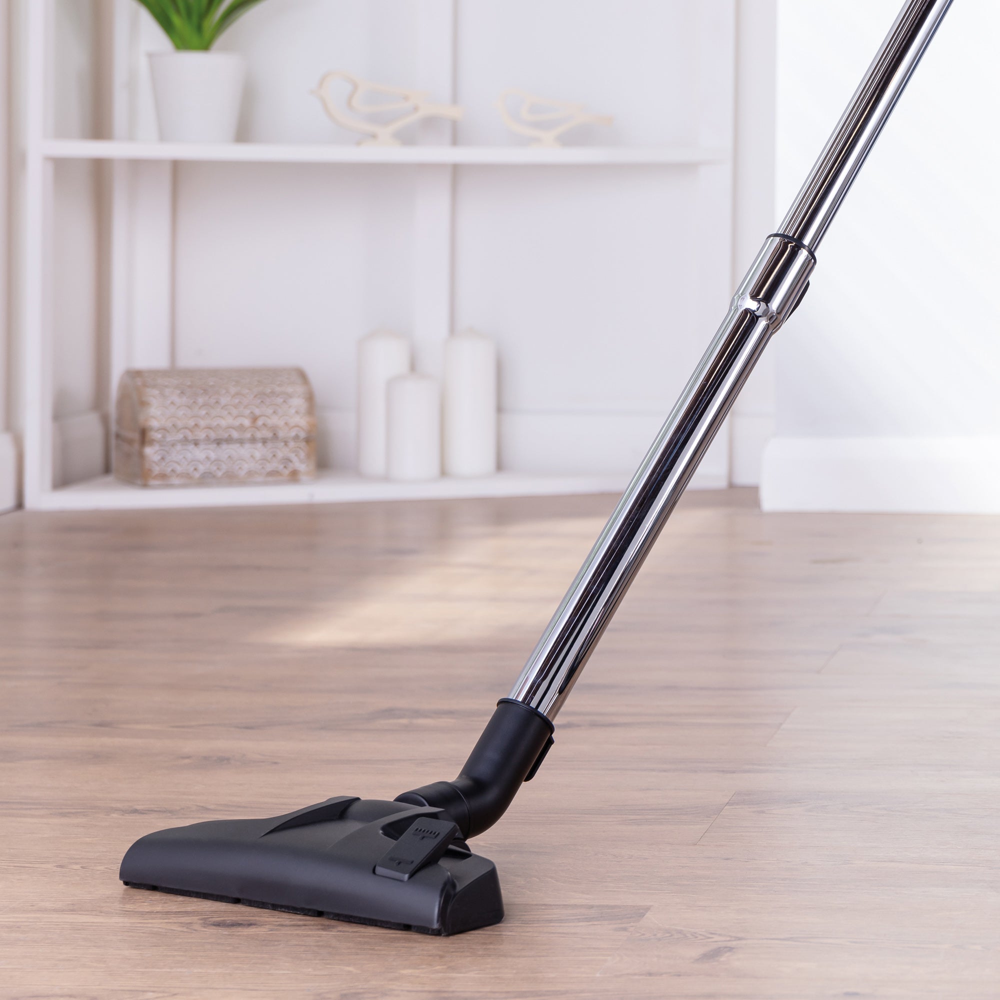 The Ewbank MOTIONLITE Bagless Cylinder Vacuum Cleaner with a metal handle and black nozzle is used to clean a wooden floor in a bright room featuring white shelves and decorative items in the background.