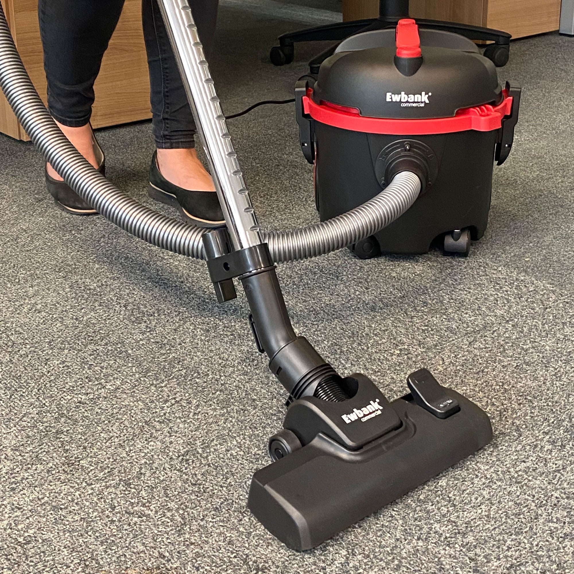 A person uses a Ewbank DV6 Dry Drum Vacuum Cleaner, black and red, with a large dust container on a grey carpet. Only their lower legs and feet in black shoes are visible. The vacuum hose connects to the wheeled canister unit.