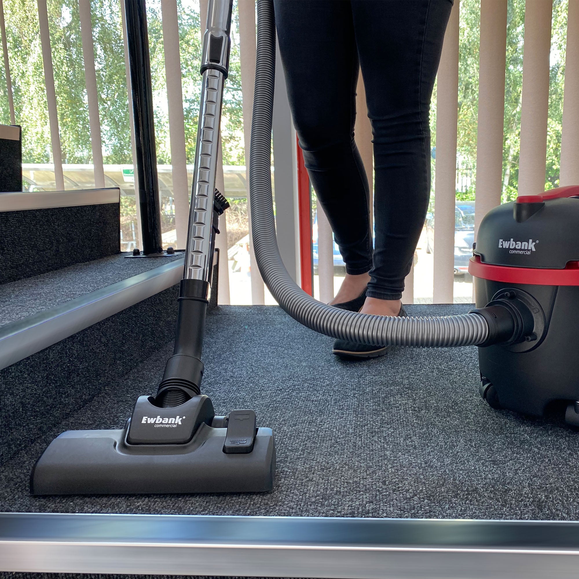 A person vacuums carpeted stairs with the DV6 Dry Drum Vacuum Cleaner, equipped with a high-efficiency floor brush, as sunlight streams through vertical blinds in the background.