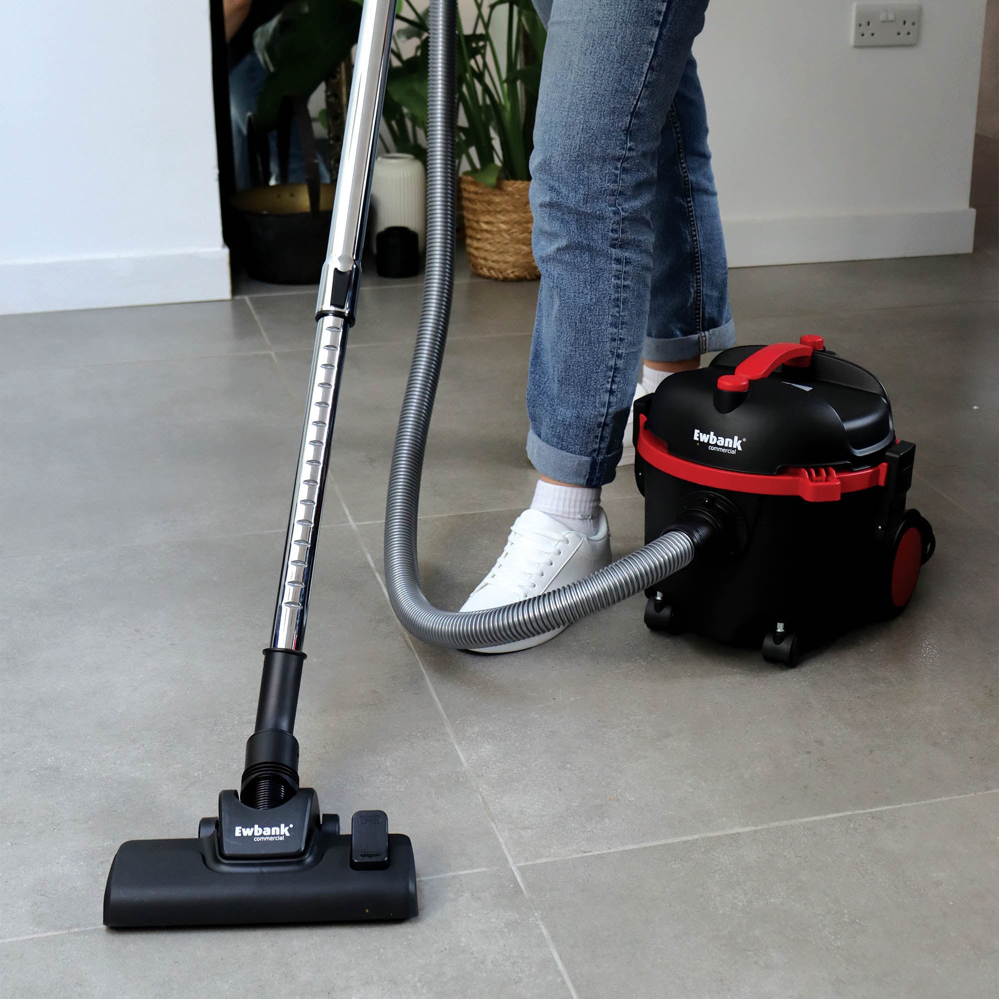 A person in jeans and white sneakers vacuums a tiled floor with the black and red DV6 Dry Drum Vacuum Cleaner featuring a high-efficiency floor brush. Green plants and a mirror are visible in the background.
