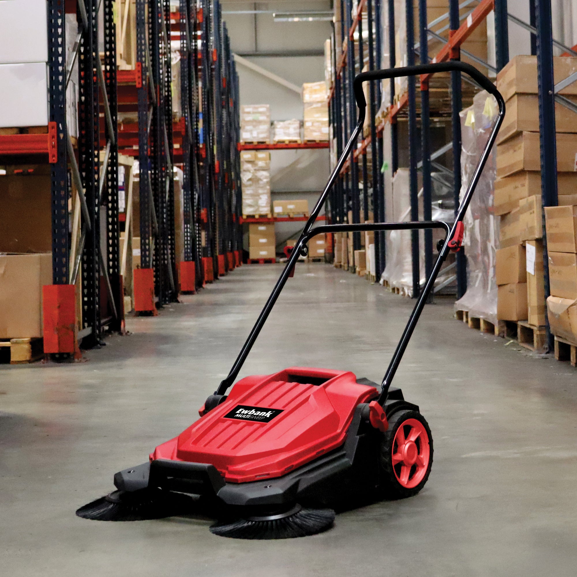 The MULTISWEEP Manual Push Sweeper, in red and black with a handle, is placed in a warehouse aisle among shelves and stacked boxes, ready for efficient outdoor cleaning.