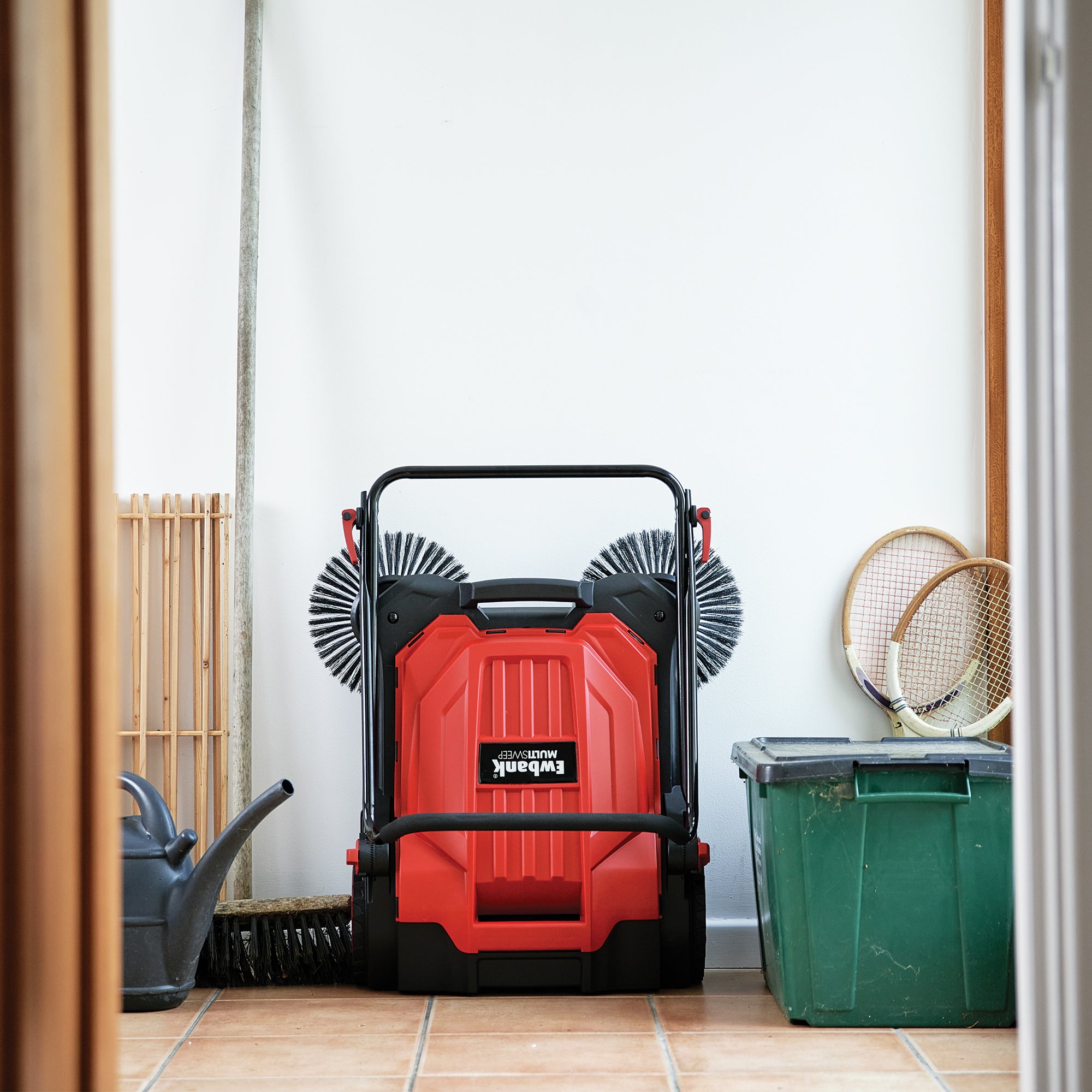 The MULTISWEEP Manual Push Sweeper in red and black stands by a white wall, next to a broom, watering can, and green storage bin with rackets—making it ideal for easy outdoor cleaning.