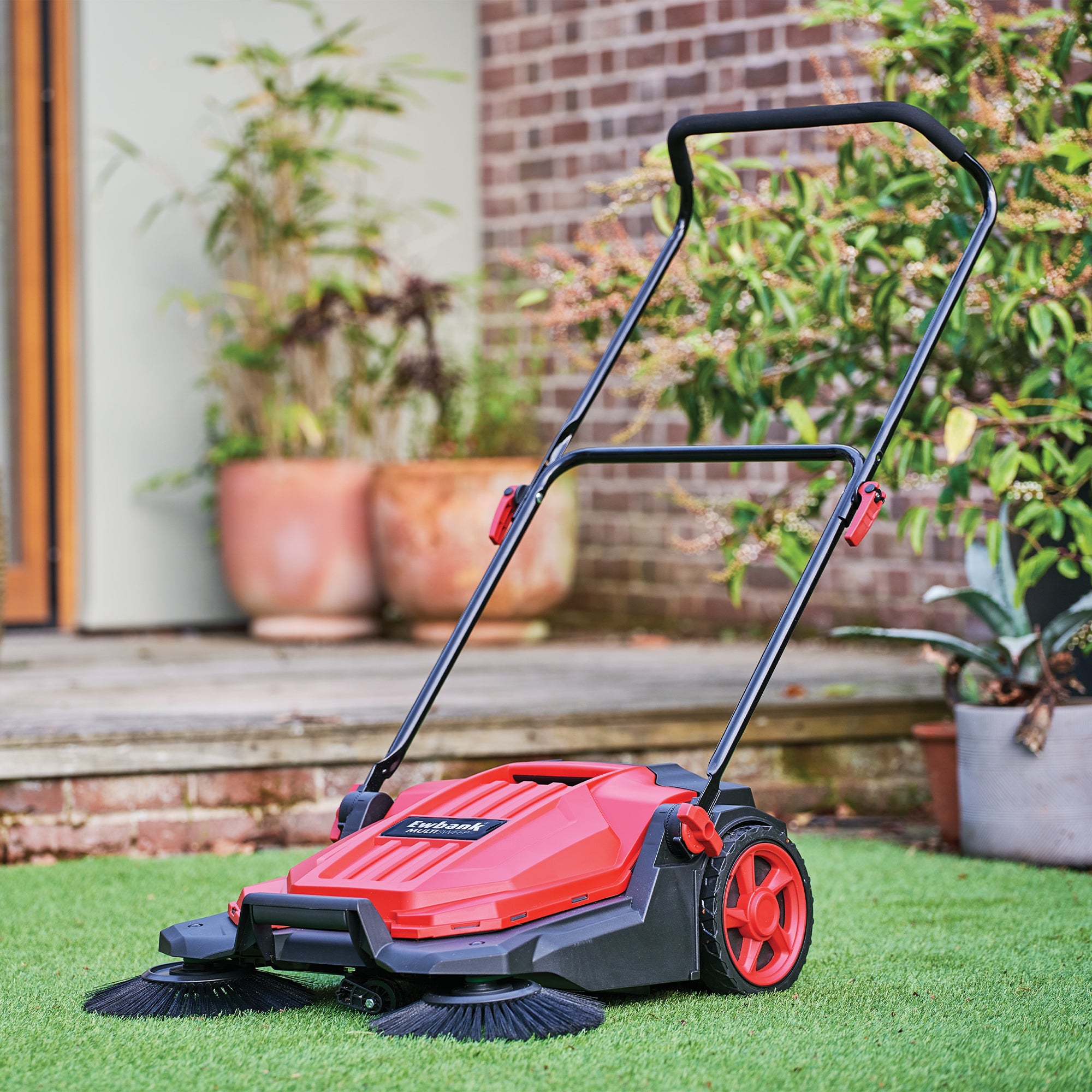 The MULTISWEEP Manual Push Sweeper in red and black, with rotating brushes, stands on artificial grass in a garden near potted plants and a brick wall, ready for outdoor cleaning.