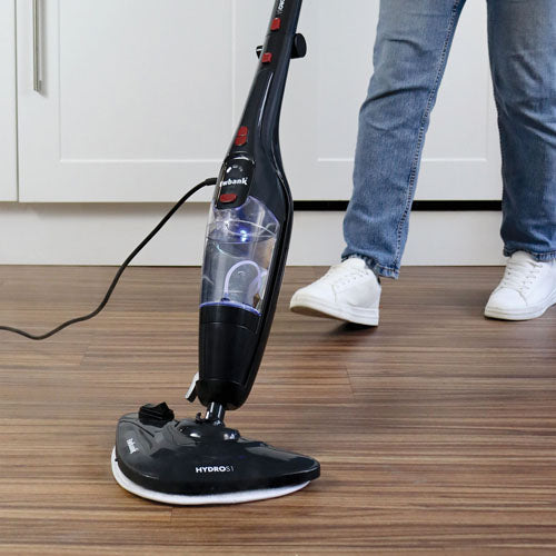 Wearing jeans and white sneakers, a person uses the Ewbank HYDROS1 5-In-1 Steam Mop in black to clean a wooden kitchen floor.