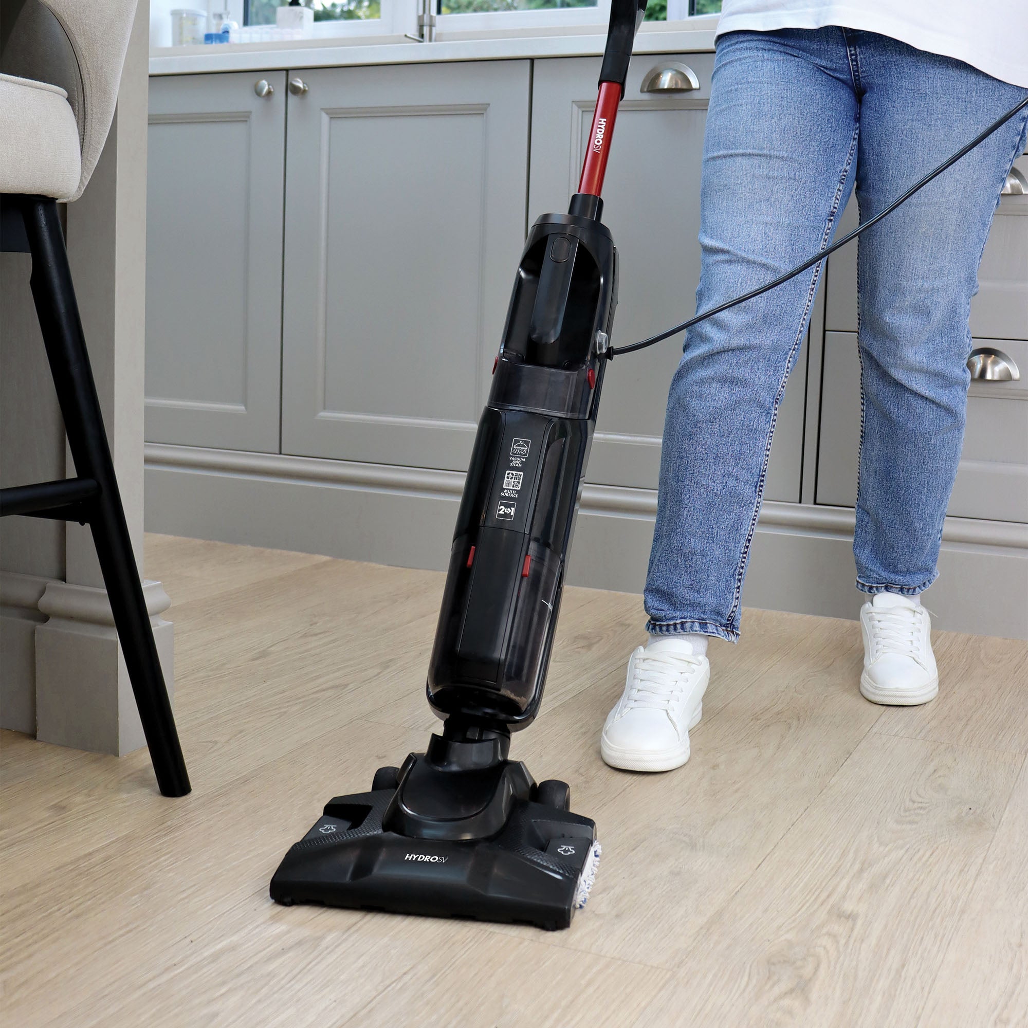 Wearing blue jeans and white sneakers, a person uses the black Ewbank HYDROSV 2-In-1 Steam Vacuum Cleaner to clean a light wood floor in a modern kitchen with sleek gray cabinets.