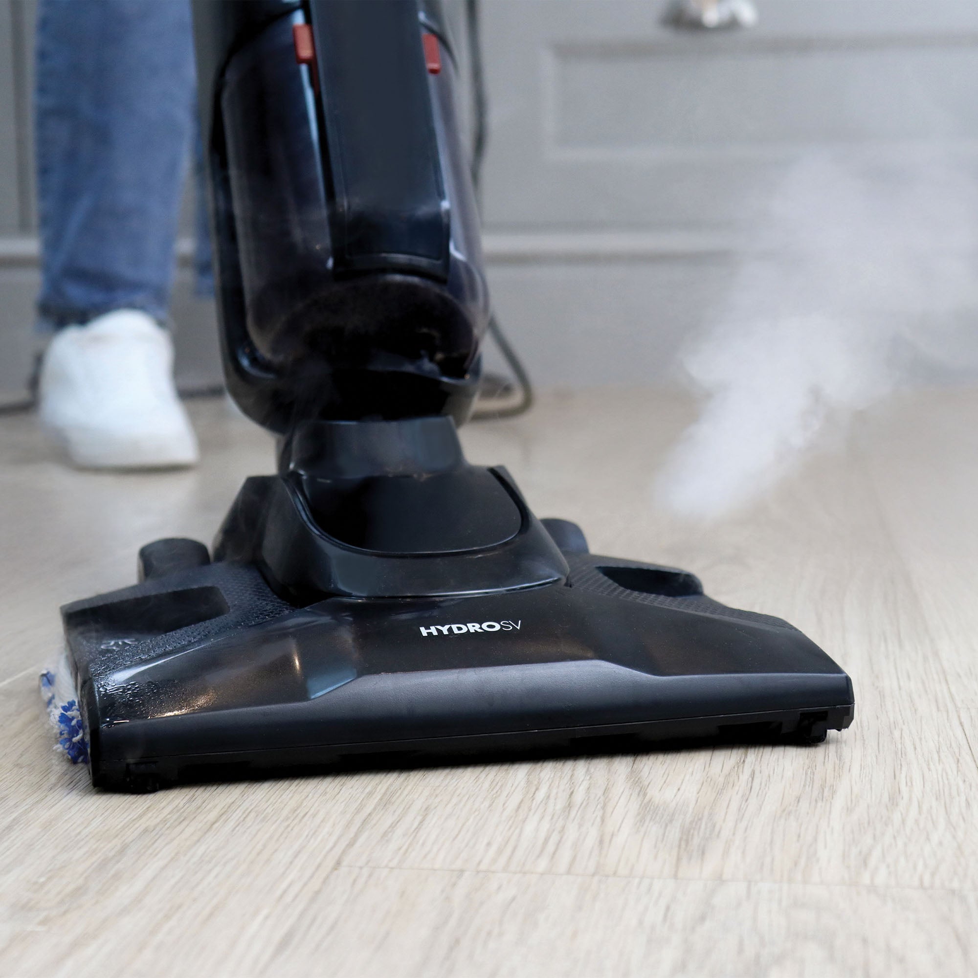 A person uses the black Ewbank HYDROSV 2-In-1 Steam Vacuum Cleaner on a light wood floor as steam is visibly emitted from the cleaner's base.
