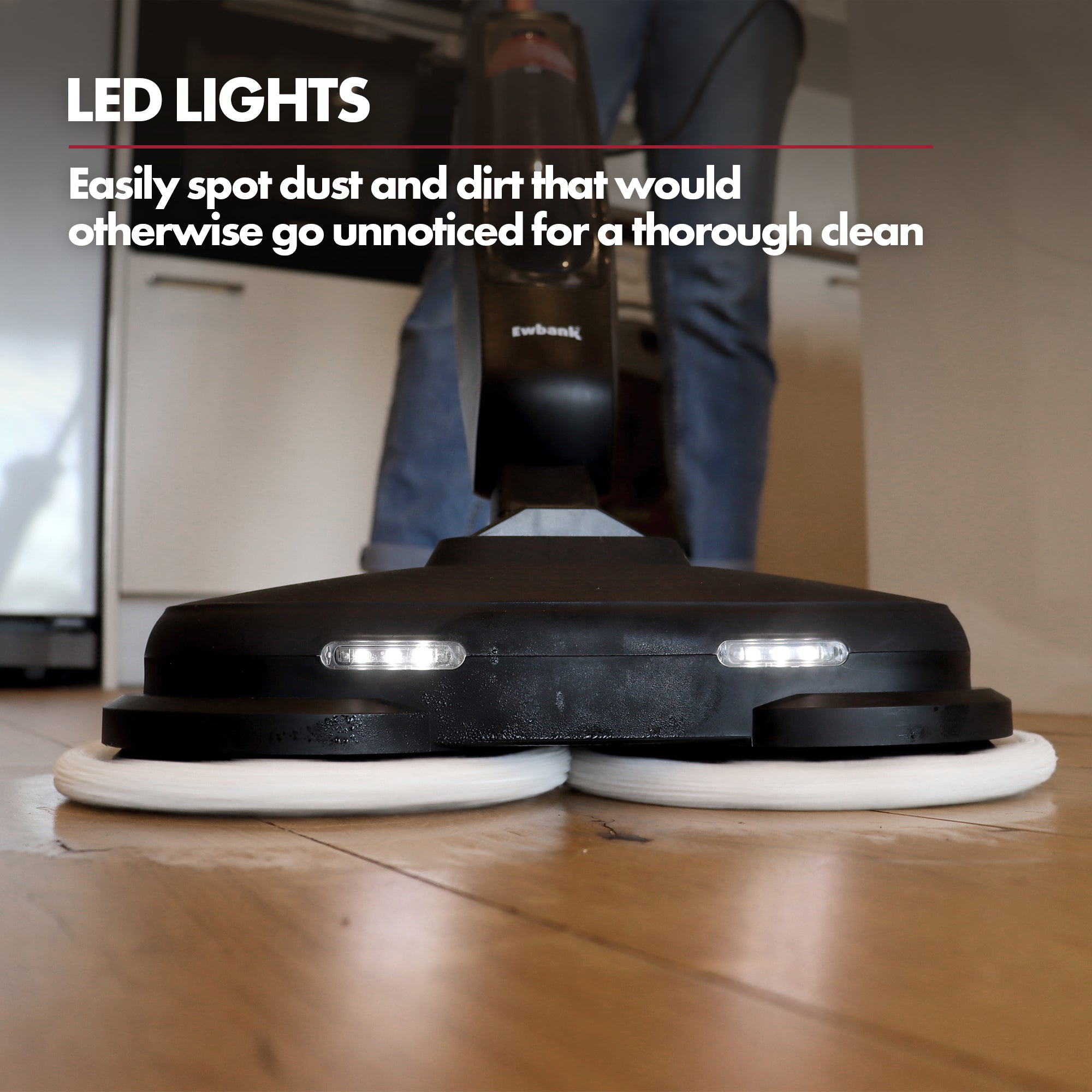 A close-up of the Ewbank EWST04079 HYDROS2 Steam and Spin Mop (1500W, 220RPM, LED lights) illuminating dust on a wooden kitchen floor, with white cabinets and a person’s legs in the background.