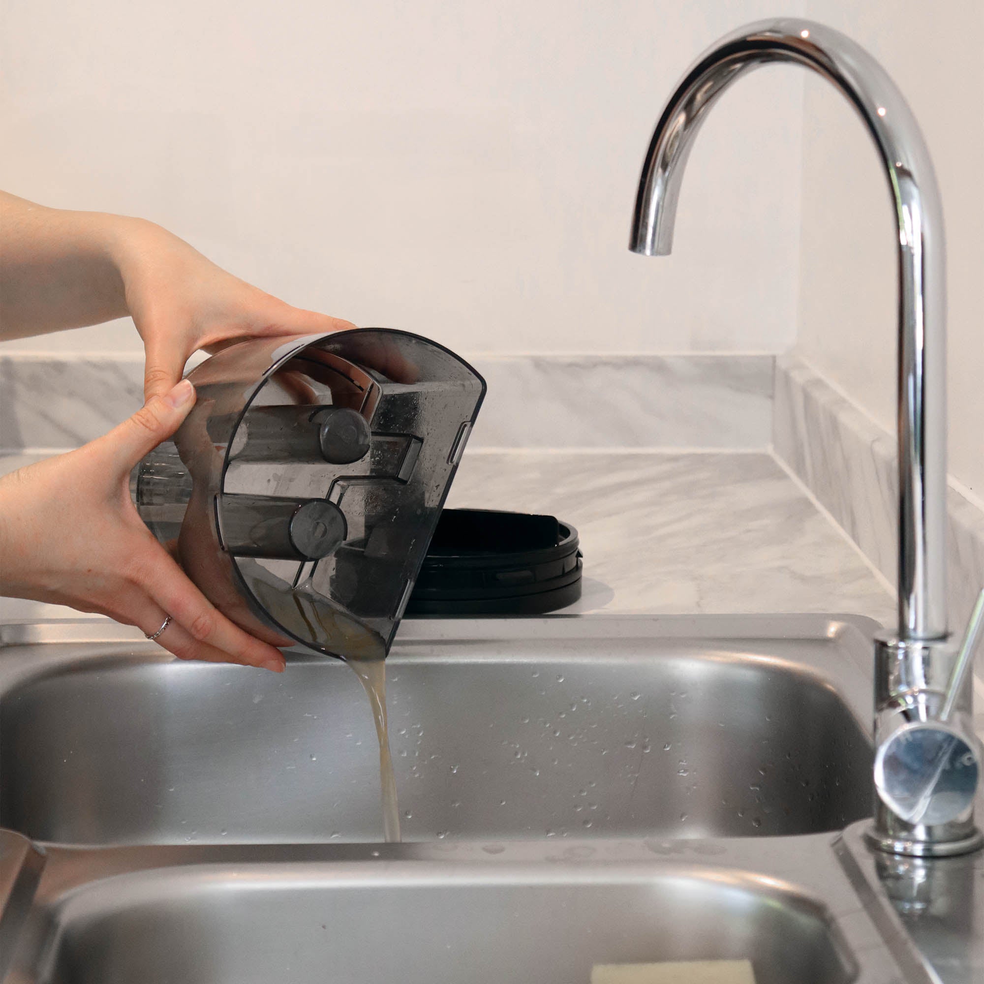 Someone is pouring dirty liquid from the HYDROSPOT Compact Spot Cleaner’s jug into a kitchen sink beside a modern chrome faucet.