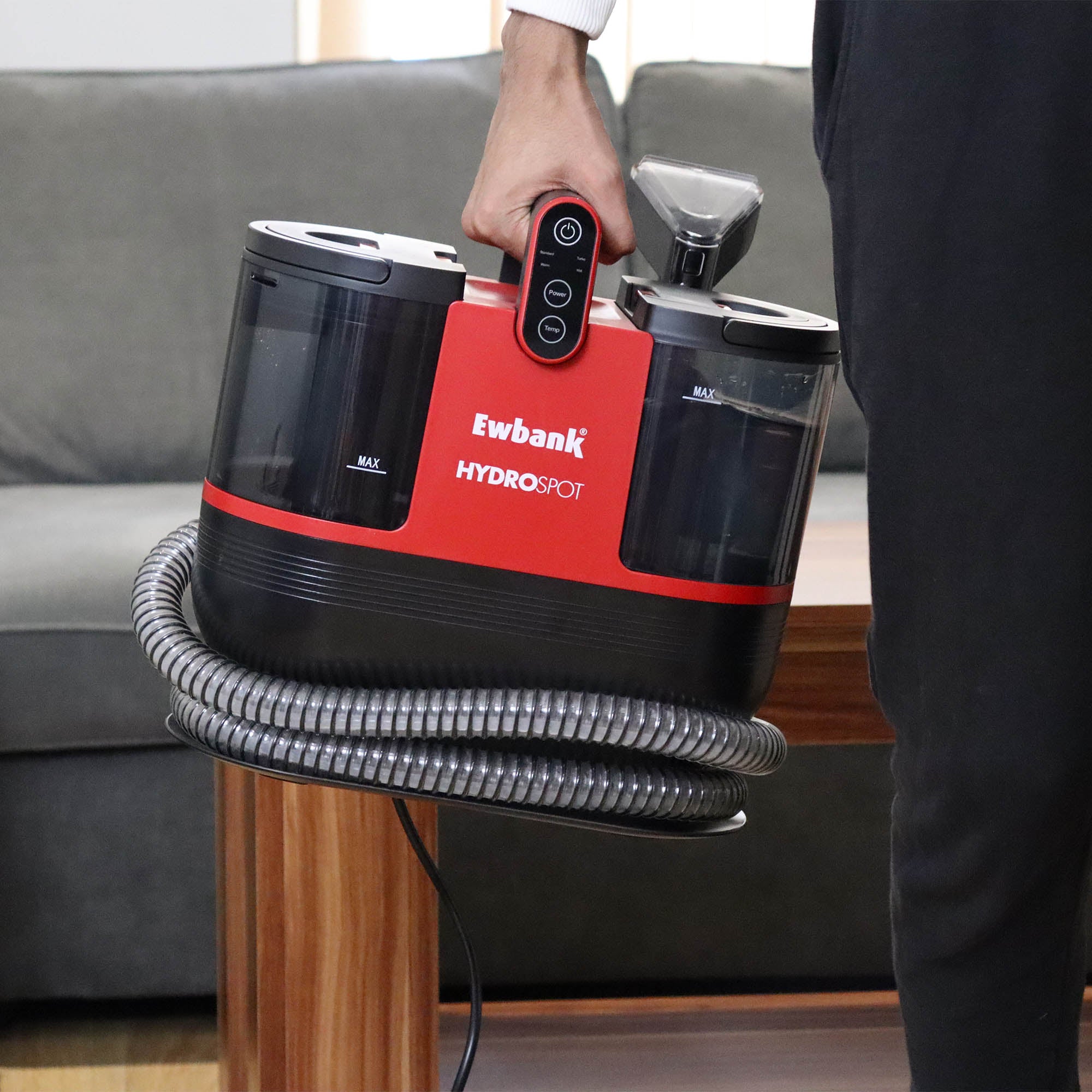 A person holds the HYDROSPOT Compact Spot Cleaner by its handle. The black and red device has clear water tanks and a coiled hose, and is shown indoors near a wooden table and gray sofa.