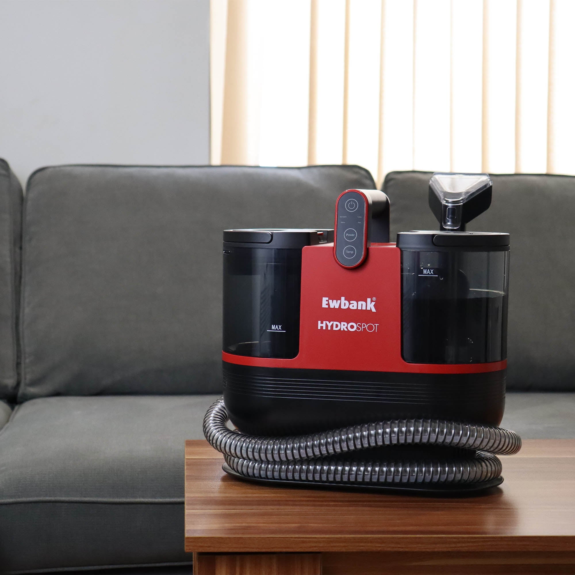 A HYDROSPOT Compact Spot Cleaner in red and black rests on a wooden table before a gray sofa, its flexible hose coiled at the base, with vertical window blinds visible in the background.