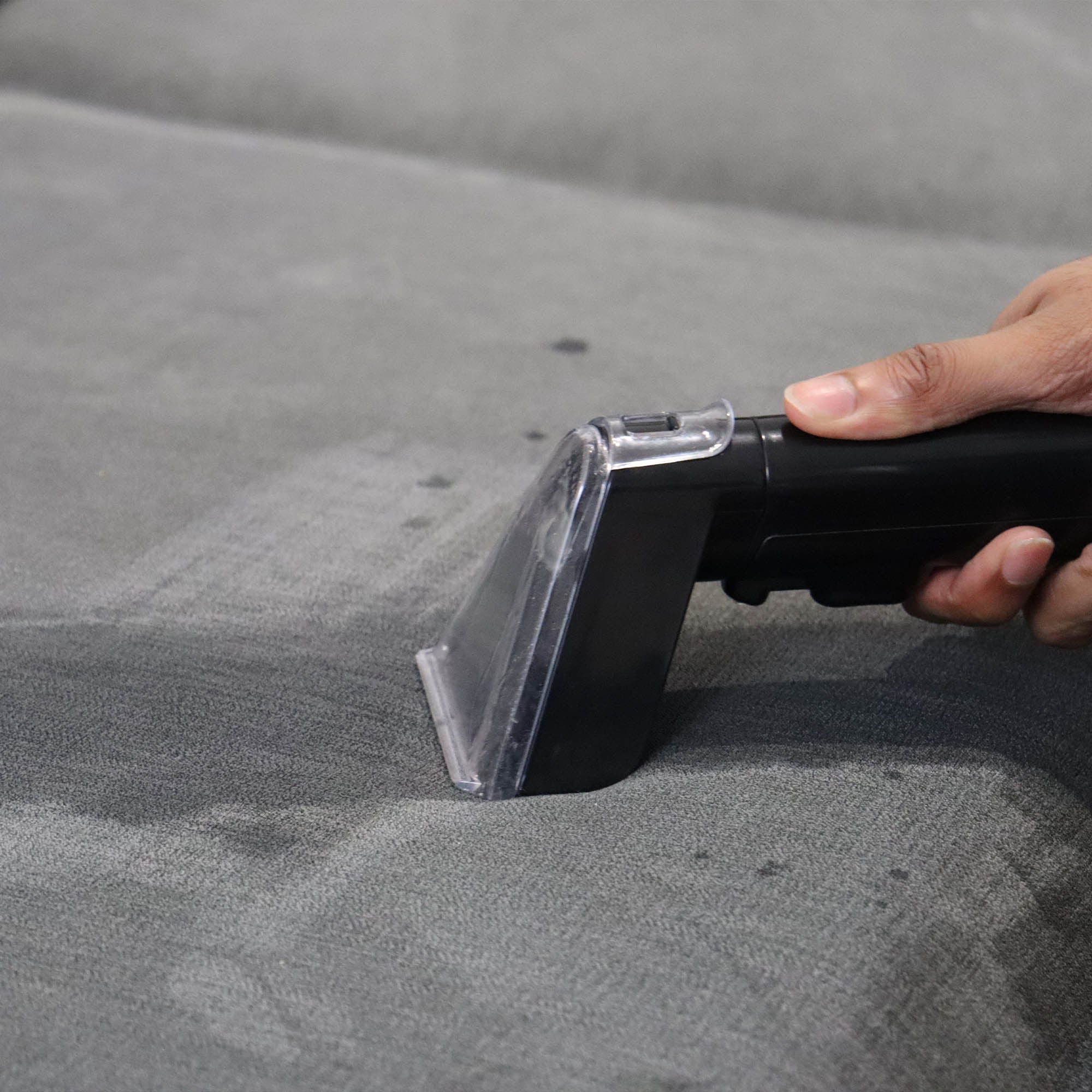 A hand uses the HYDROSPOT Compact Spot Cleaner to clean a gray fabric surface, removing visible dirt spots and revealing freshly cleaned areas.